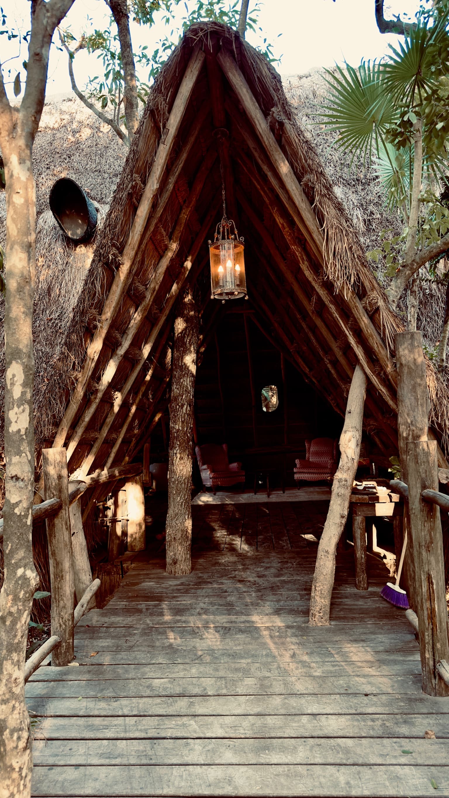 A wooden path leading to an A-framed hut with a straw roof, hanging lantern and trees in the surrounding area.