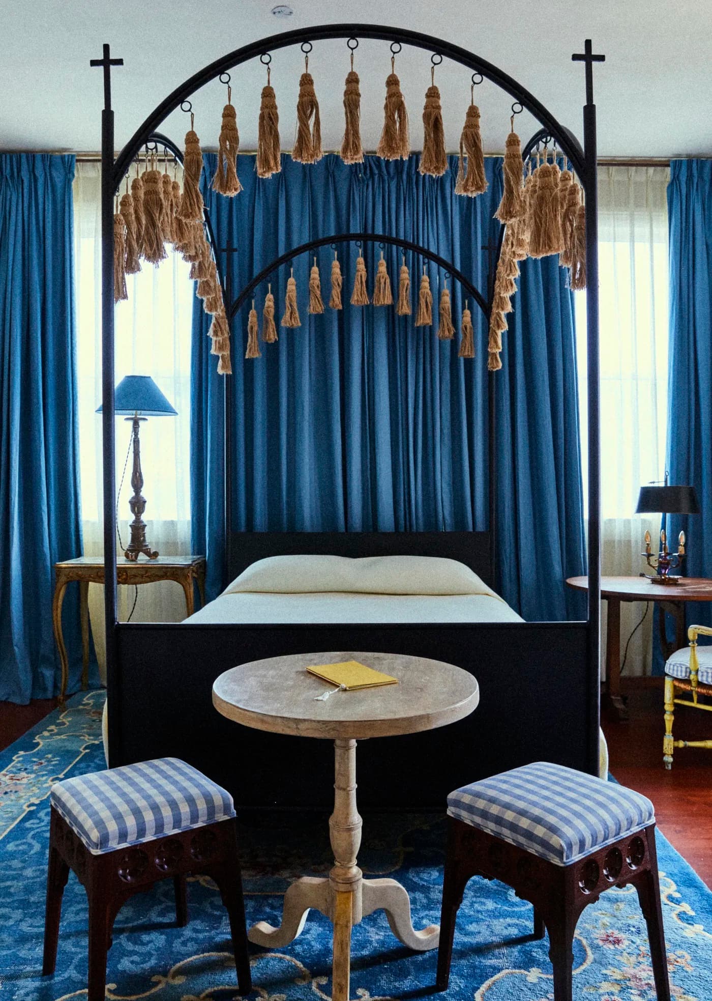 Image 1: A master bedroom, with a king-sized bed, wooden canopy topped with crosses, royal blue curtains, and a small round table and stools.