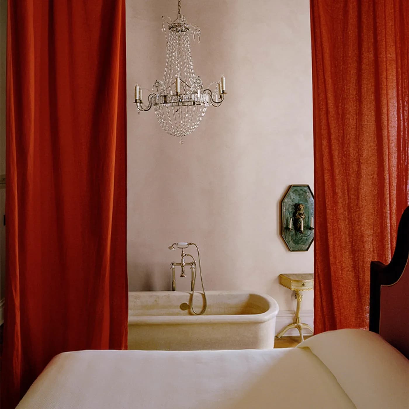 A bed with a white blanket in front of ceiling-high, red curtains, revealing a bathtub in the background.