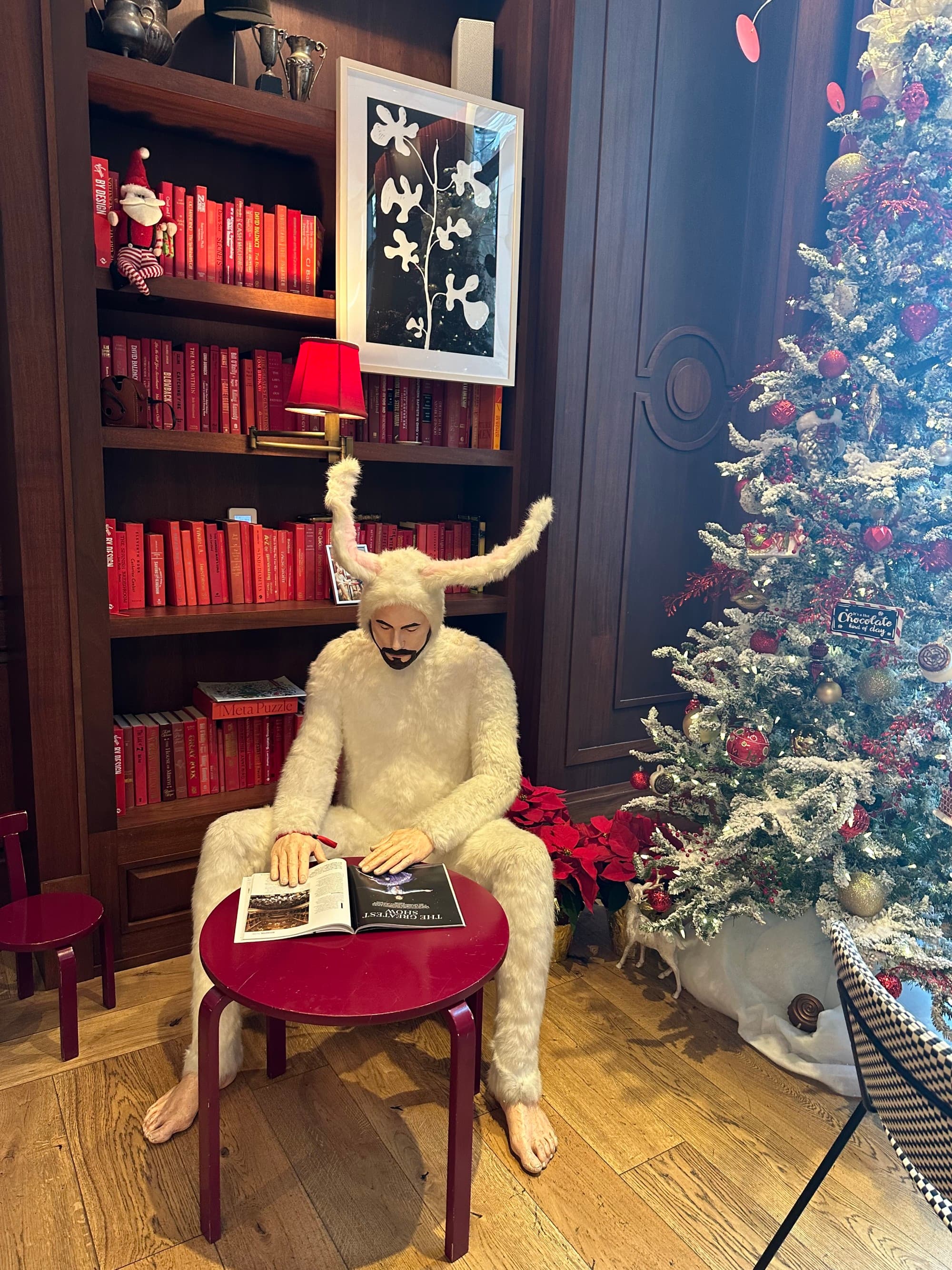 Unique artwork at the hotel of a man dressed in a furry bunny outfit sitting at a small red table reading. There is a bookshelf of red books behind and a christmas tree in view.