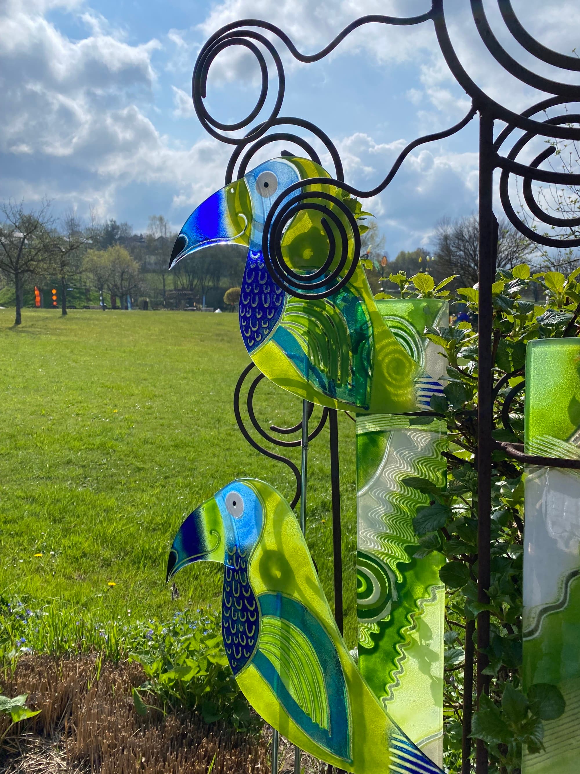 Lawn ornaments of two green and blue parrots, planted in a vast, green field on a sunny day.