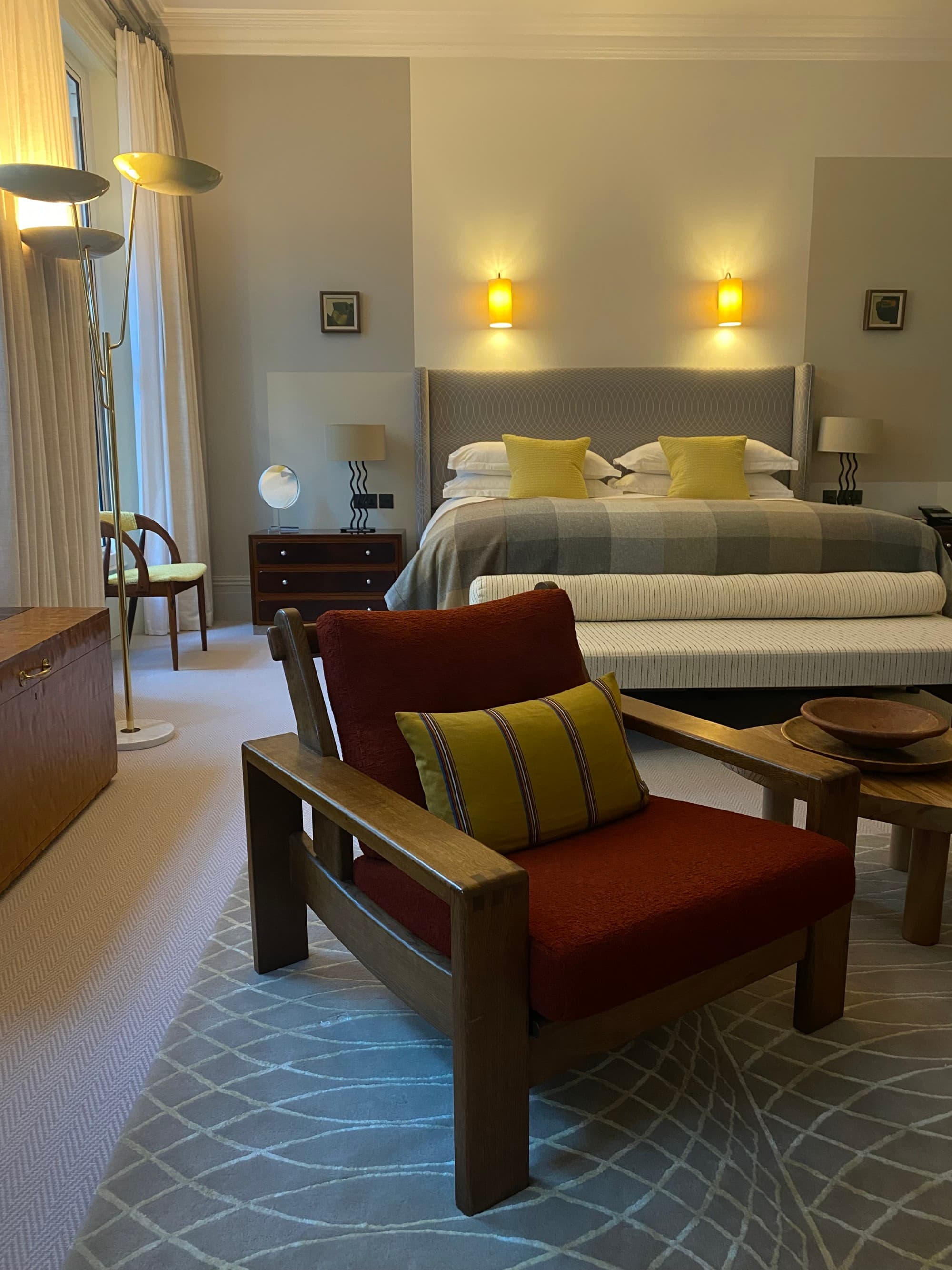 A photo inside one of the hotel rooms. In it stands a large bed with gray and yellow bedding, a red lounge chair and warm lights mounted on the walls.