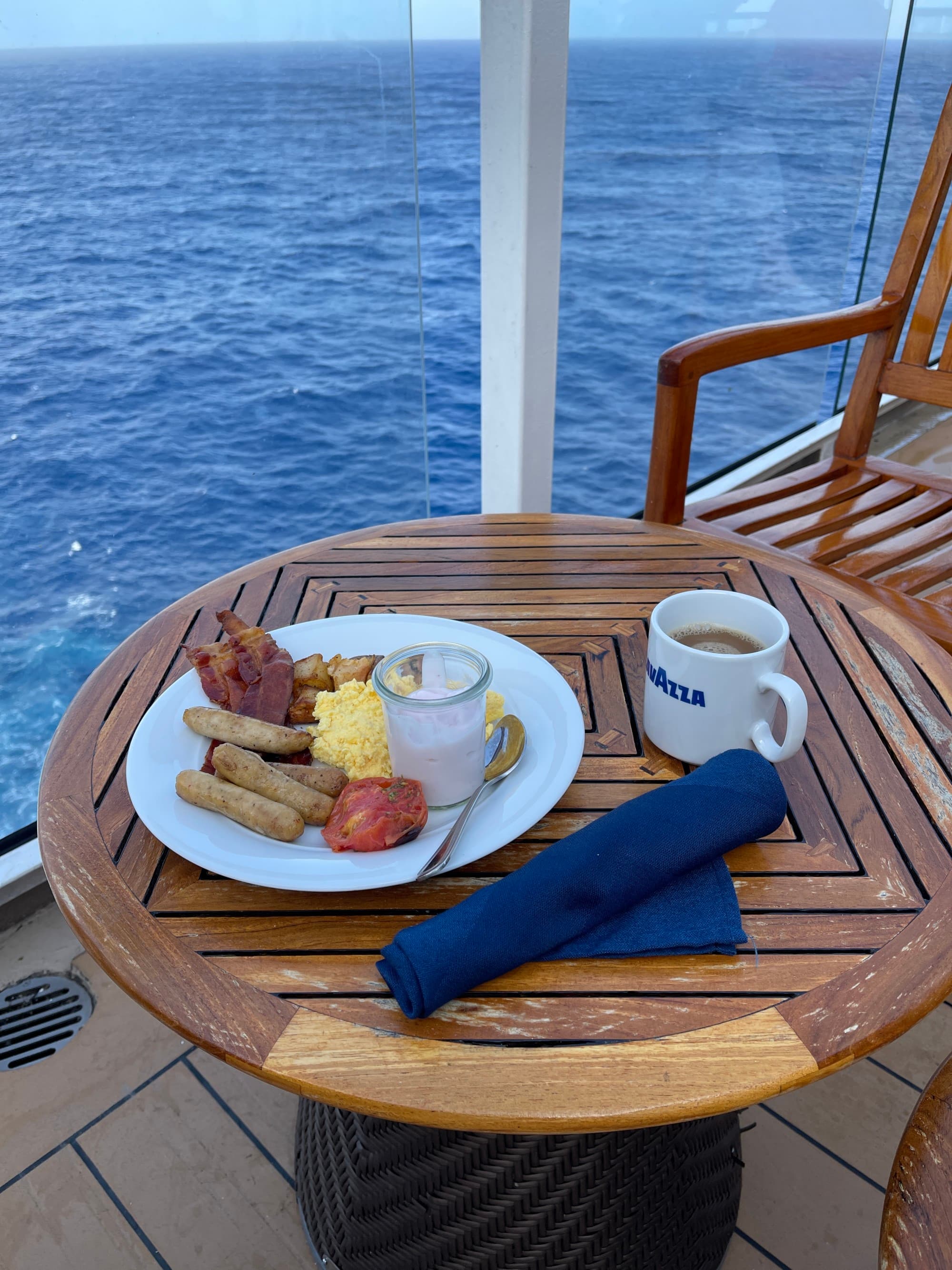 A breakfast plate of eggs, bacon, sausage and coffee on a wooden, round table, outside on the deck of the sip, with the big blue sea in the background.