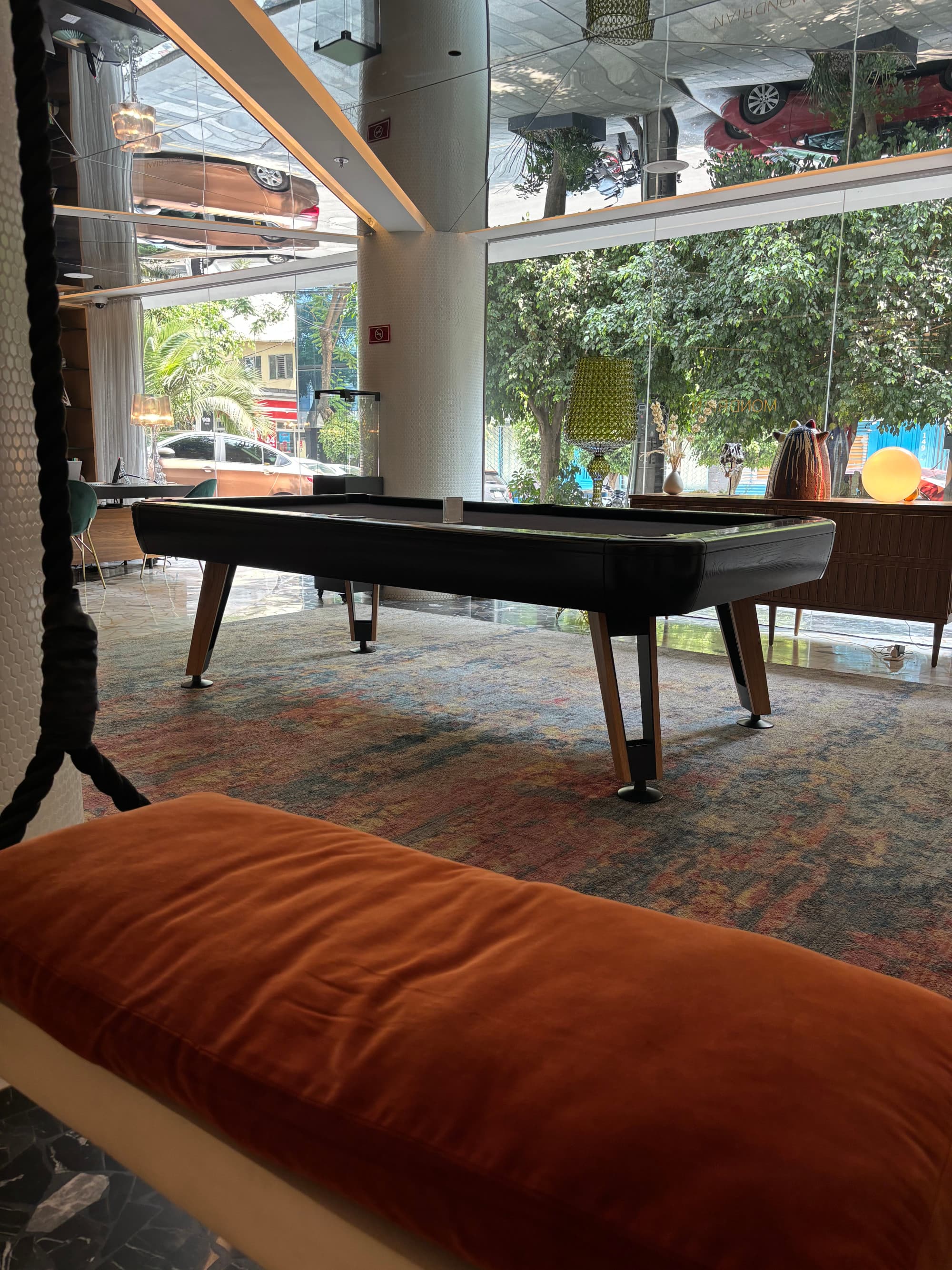 A pool table set in a lounge area for guests, with floor-ceiling windows showcasing trees and buildings outside.