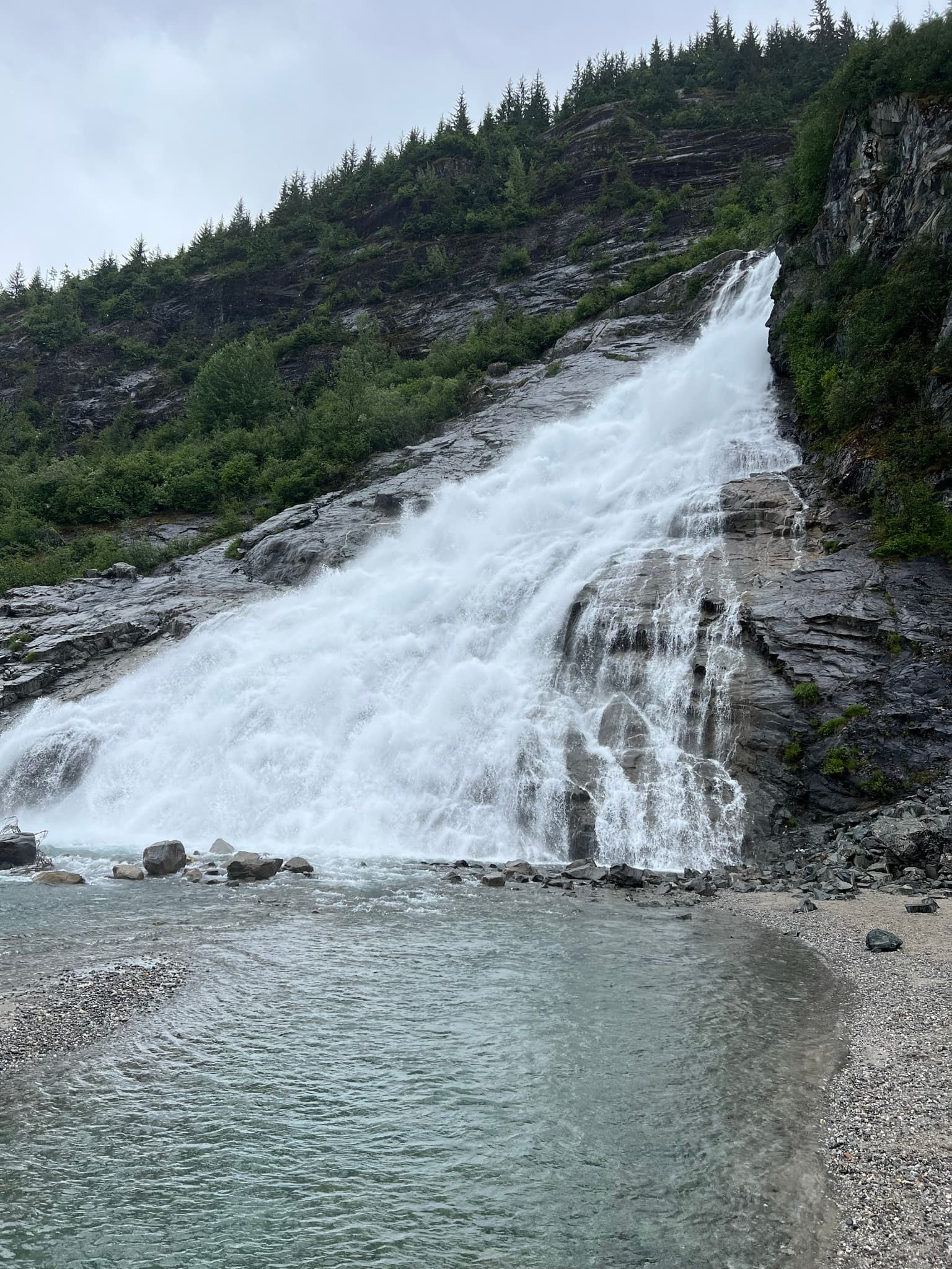 A waterfall gushing down a hill of rocky terrain, into a small pool of water among a green forest.