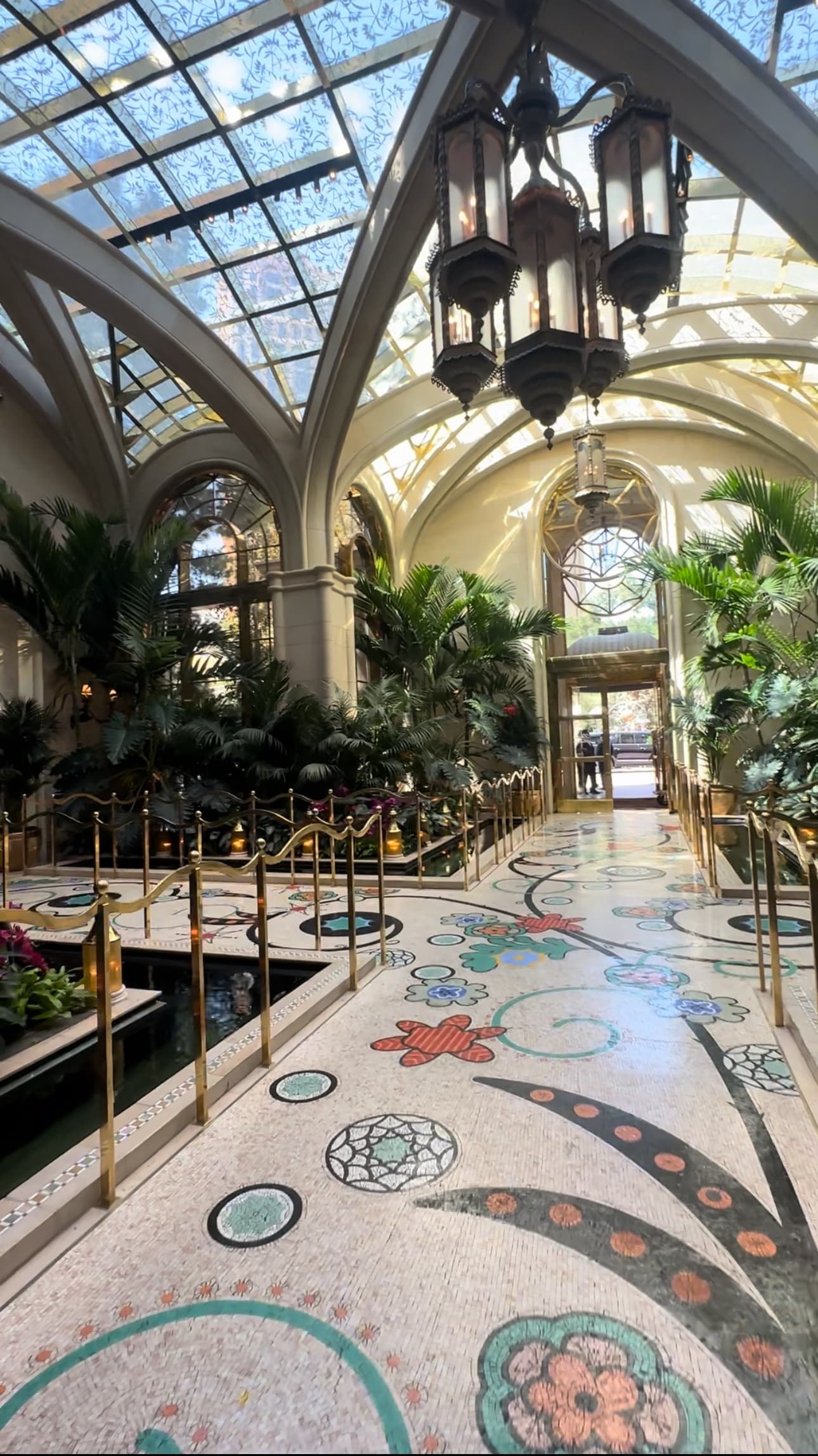 The hotel lobby with a mosaic floor, curved glass for the ceilings, a hanging lantern and trees and shrubs.