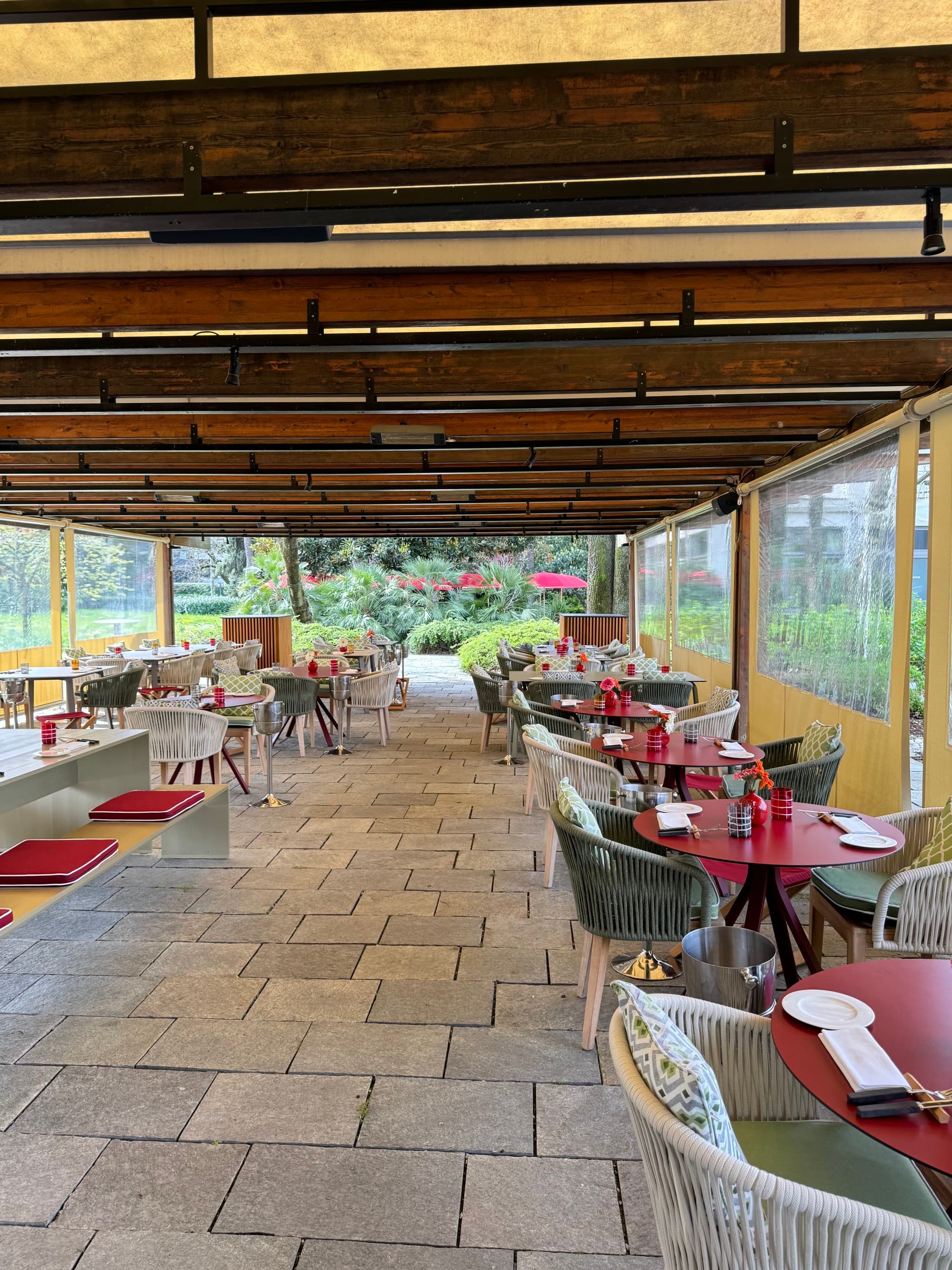 A long, covered, outdoor seating area with a stone tiled floor and a garden at the end of it.