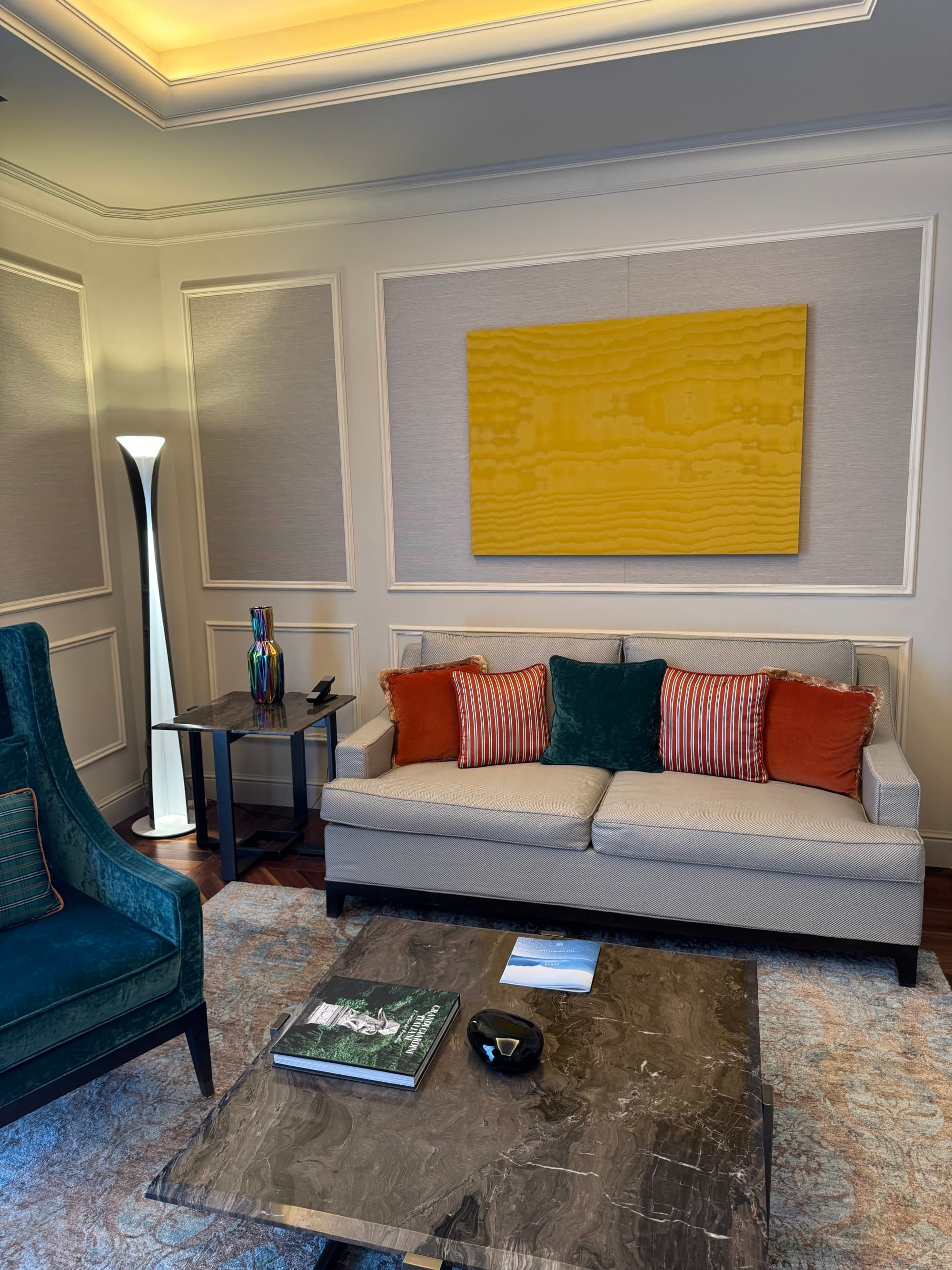 A sitting area with a couch, lounge chair and a coffee table. The wall is painted different shades of light gray with a mustard yellow painting hanging on it.