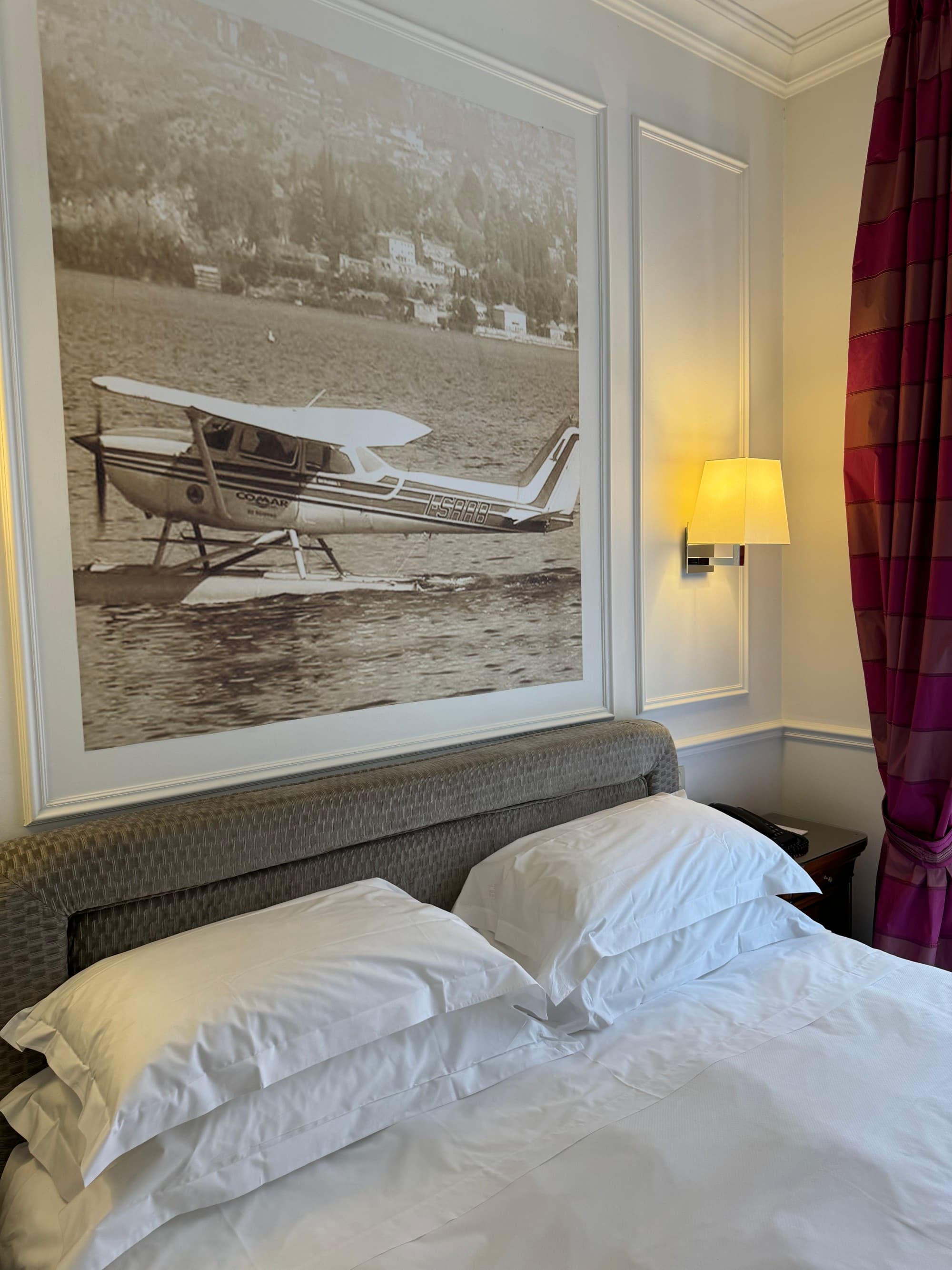 Inside a hotel room looking at a white linen bed with a gray fabric headboard and a photo of a vintage seaplane hung above it.