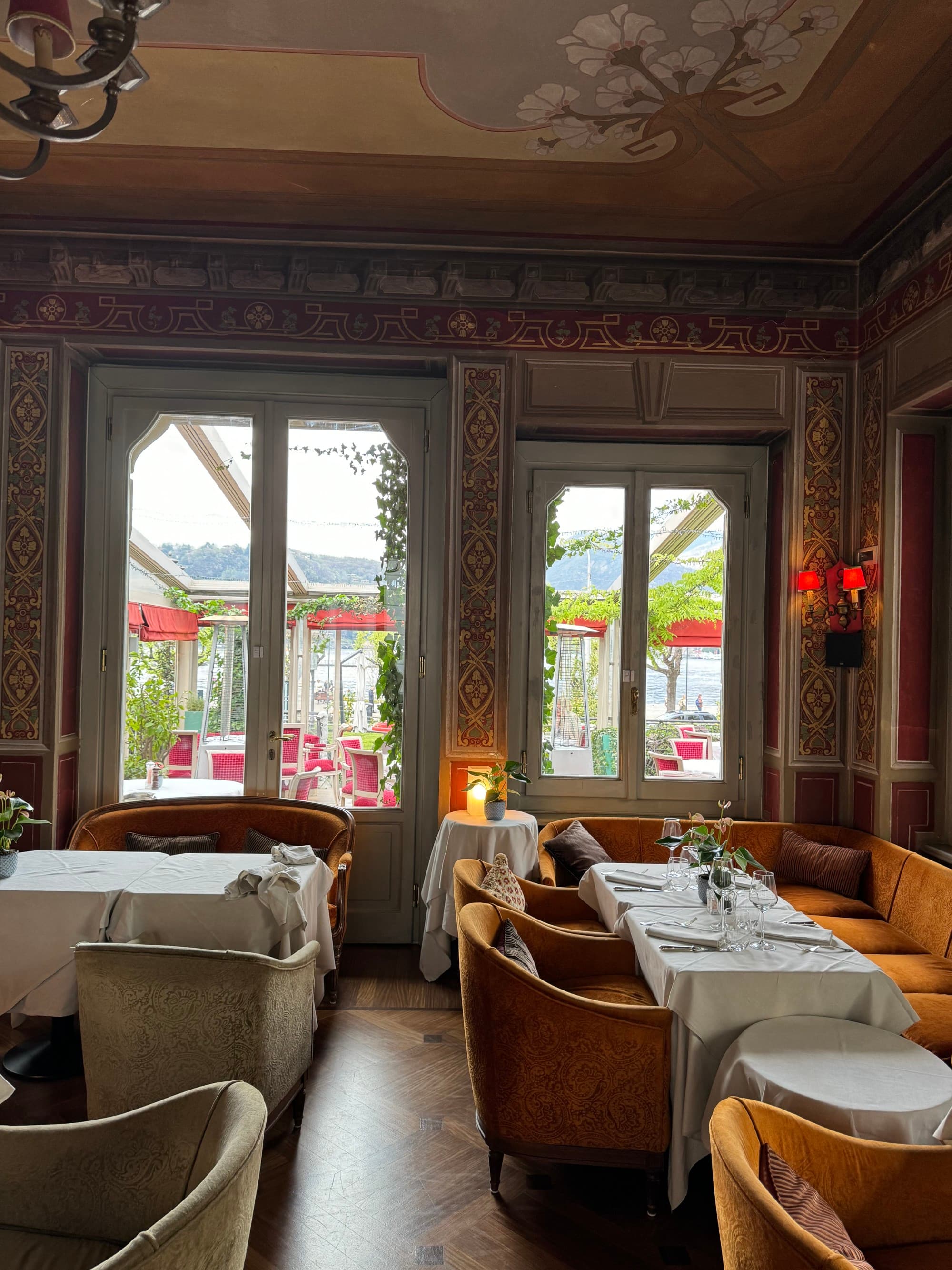 A dining room with ornate wall and ceiling paintings incorporating touches of gold. Large windows let in light on the opposite side of the room and a metal chandelier hands from the center of the ceiling.