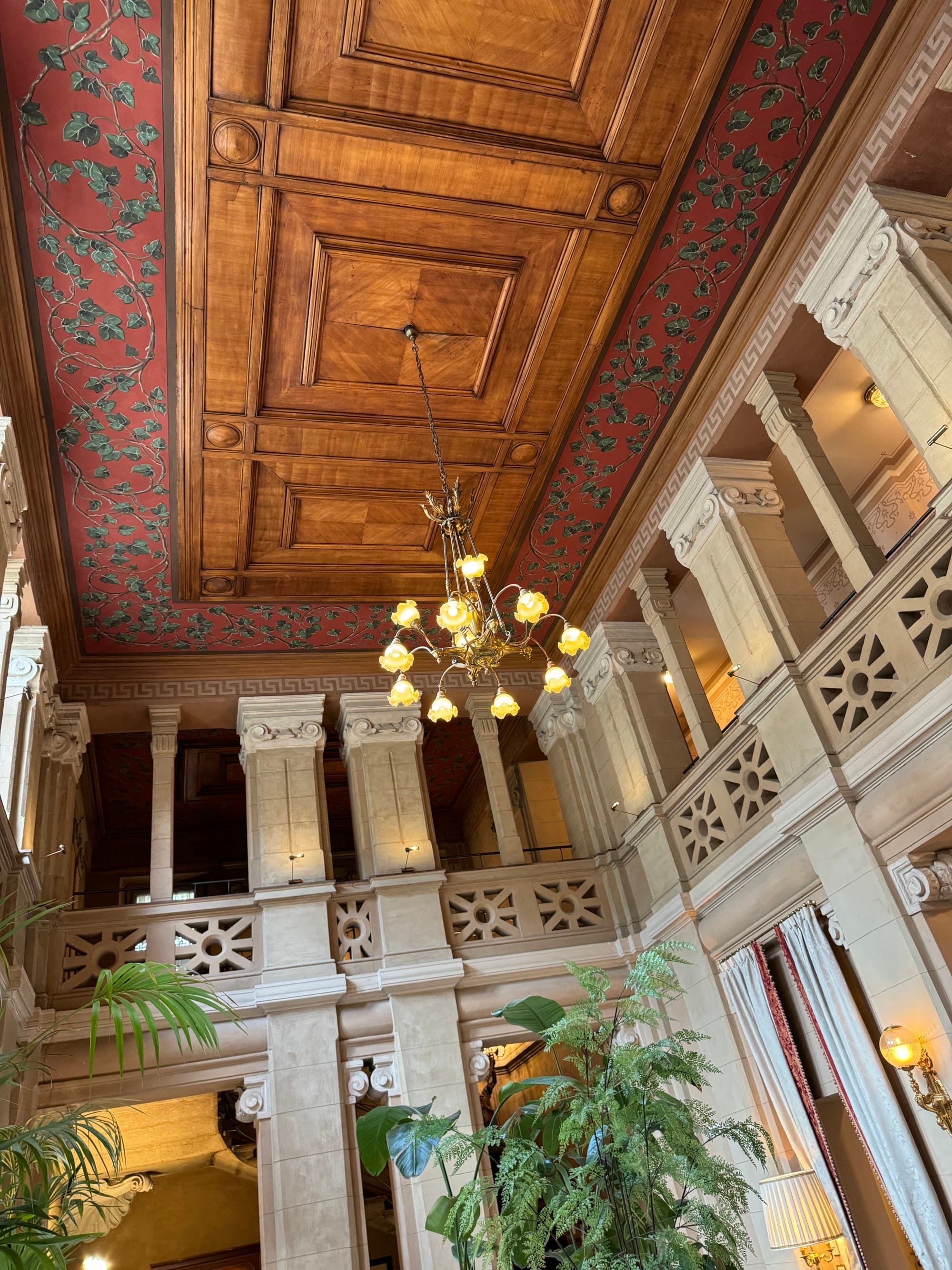 A photo of a lobby with a second-level floor balcony spanning the walls, a large chandelier hanging from the ceiling decorated with wooden paneling and red and green floral wallpaper.