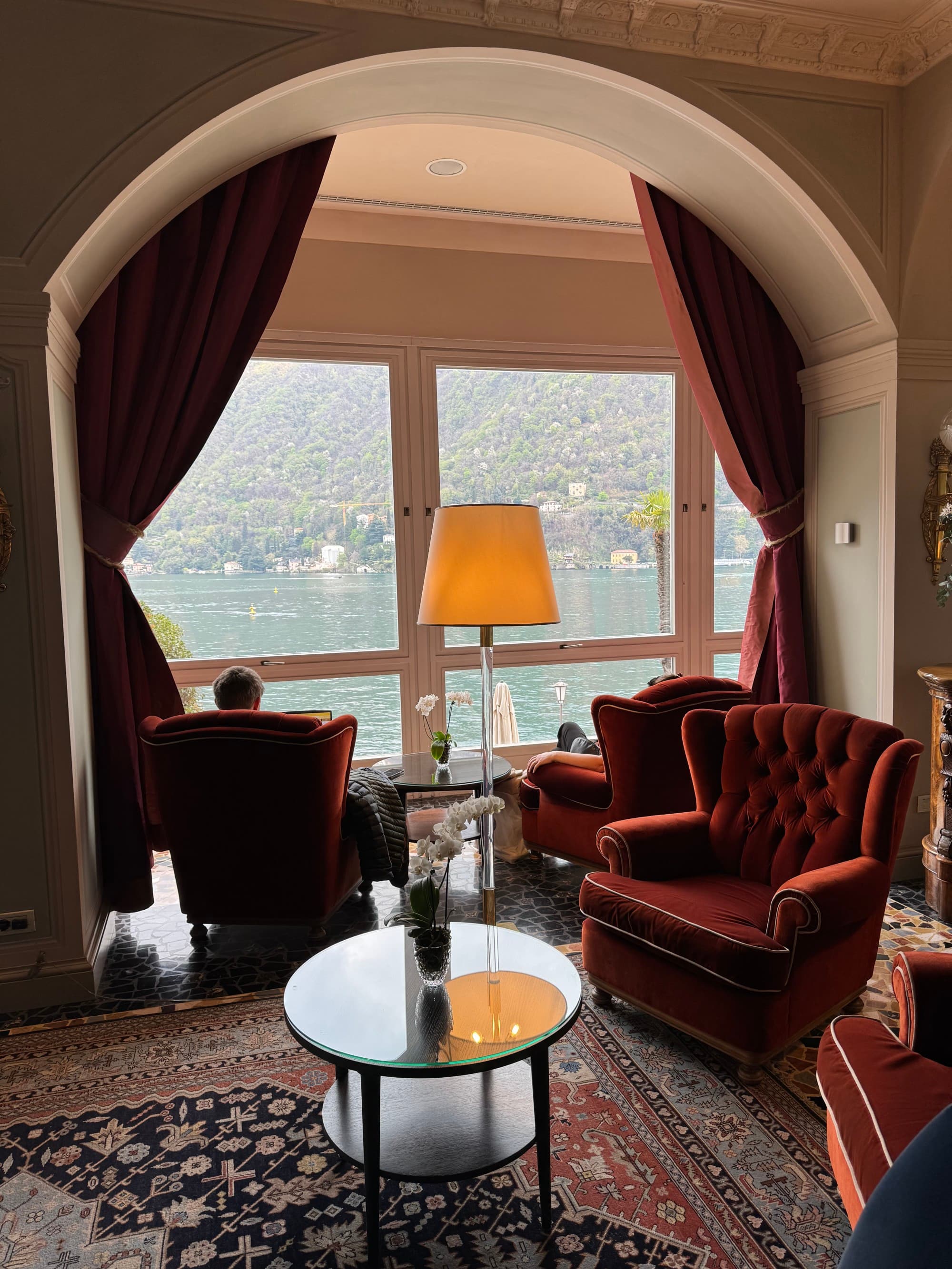 A room with indoor seating filled with large, plush red lounge chairs. Some of which sit underneath an archway next to large windows looking out over the lake.