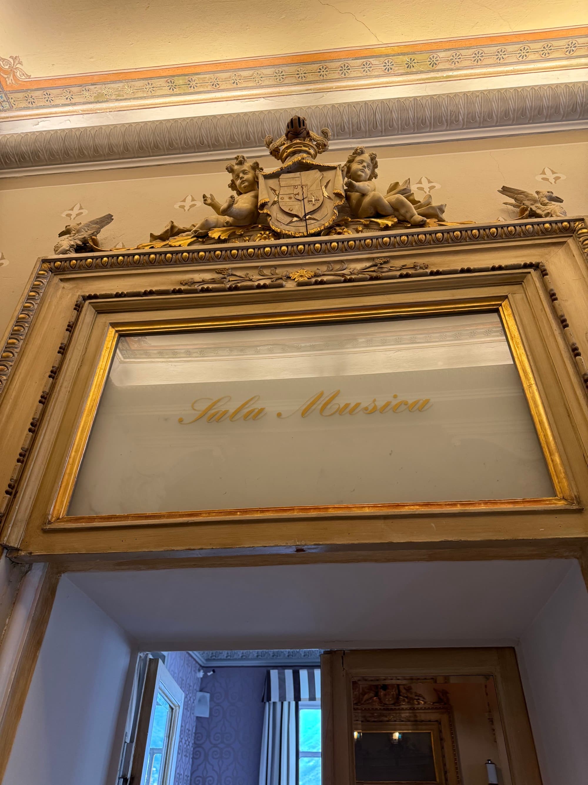 A photo of the top of an ornate, golden sculpted doorway with a painted glass sign reading " Sala Musica".