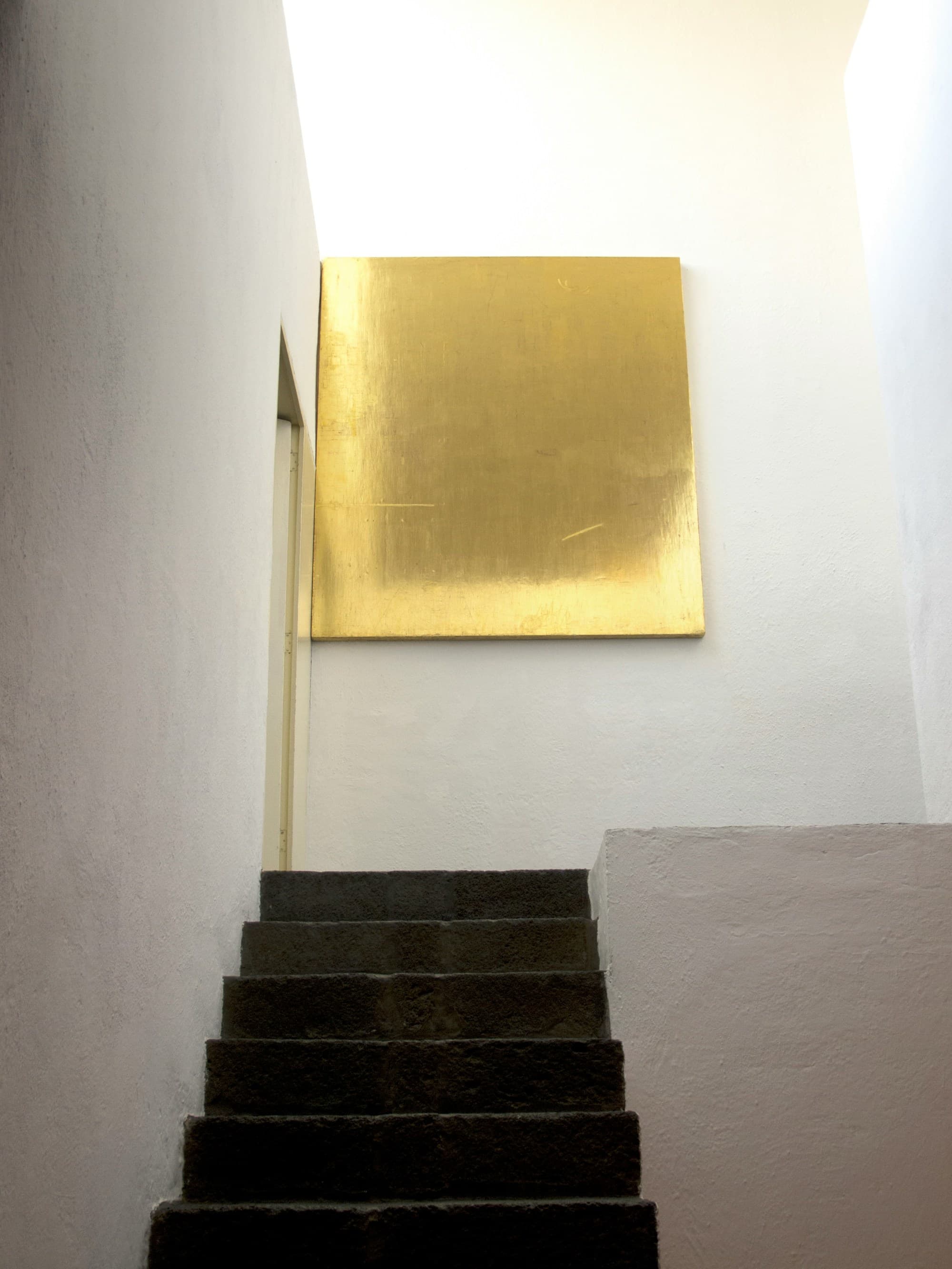 a staircase leads up to a gold square hanging on a sparse white wall