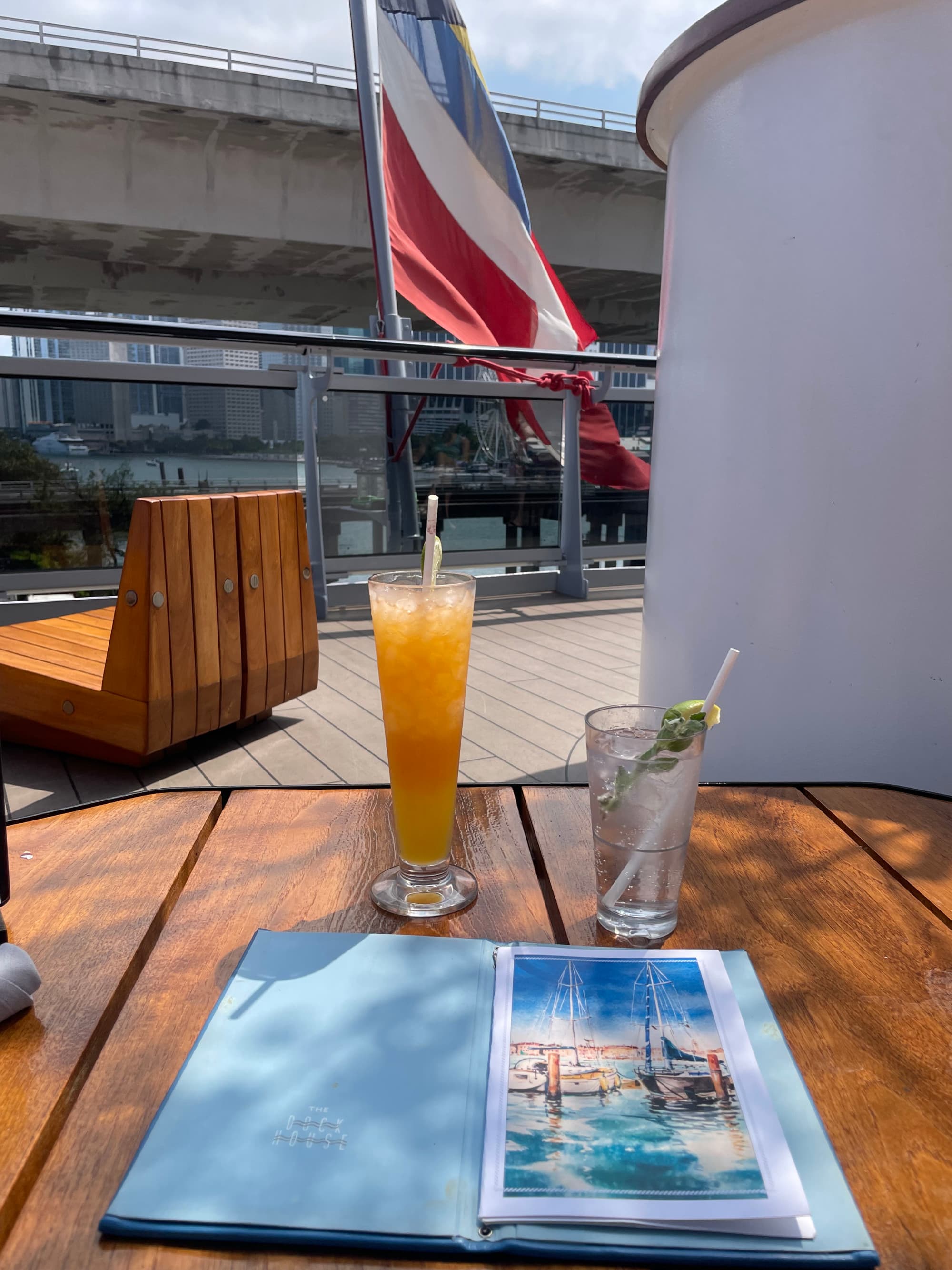 A view of a drink and a menu on a table on the deck of a cruise ship wit a flag flying behind.