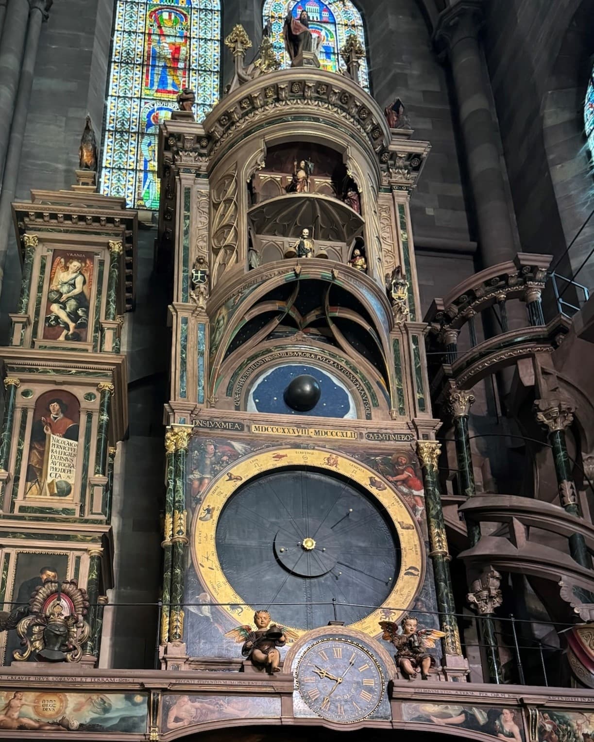 A giant bronze historical clock called the Astronomical clock at Strausburg Cathedral with stained glass windows behind it.