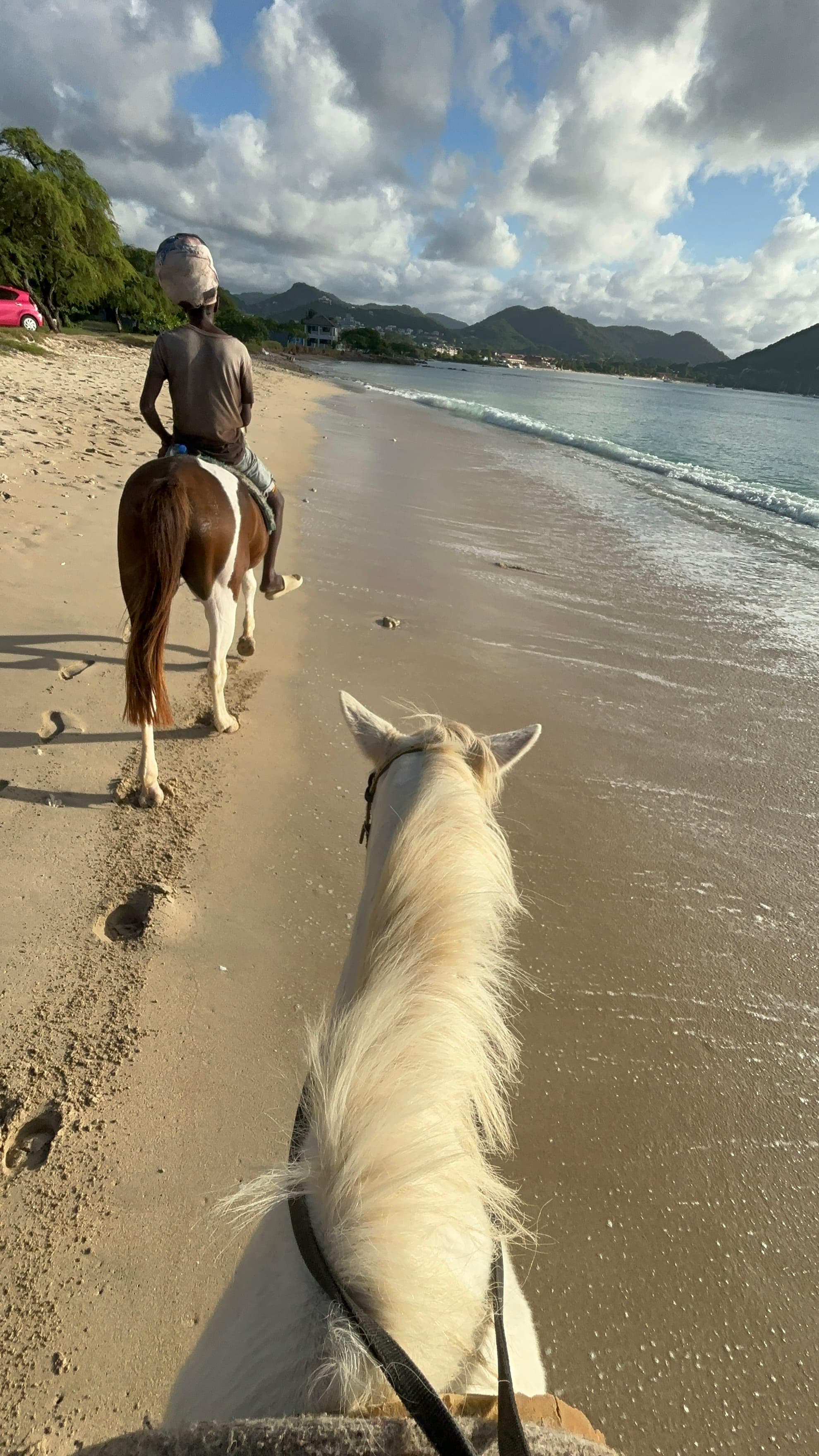 A couple on horseback clop along the beach as waves lap the shore on a sunny day dotted with clouds.