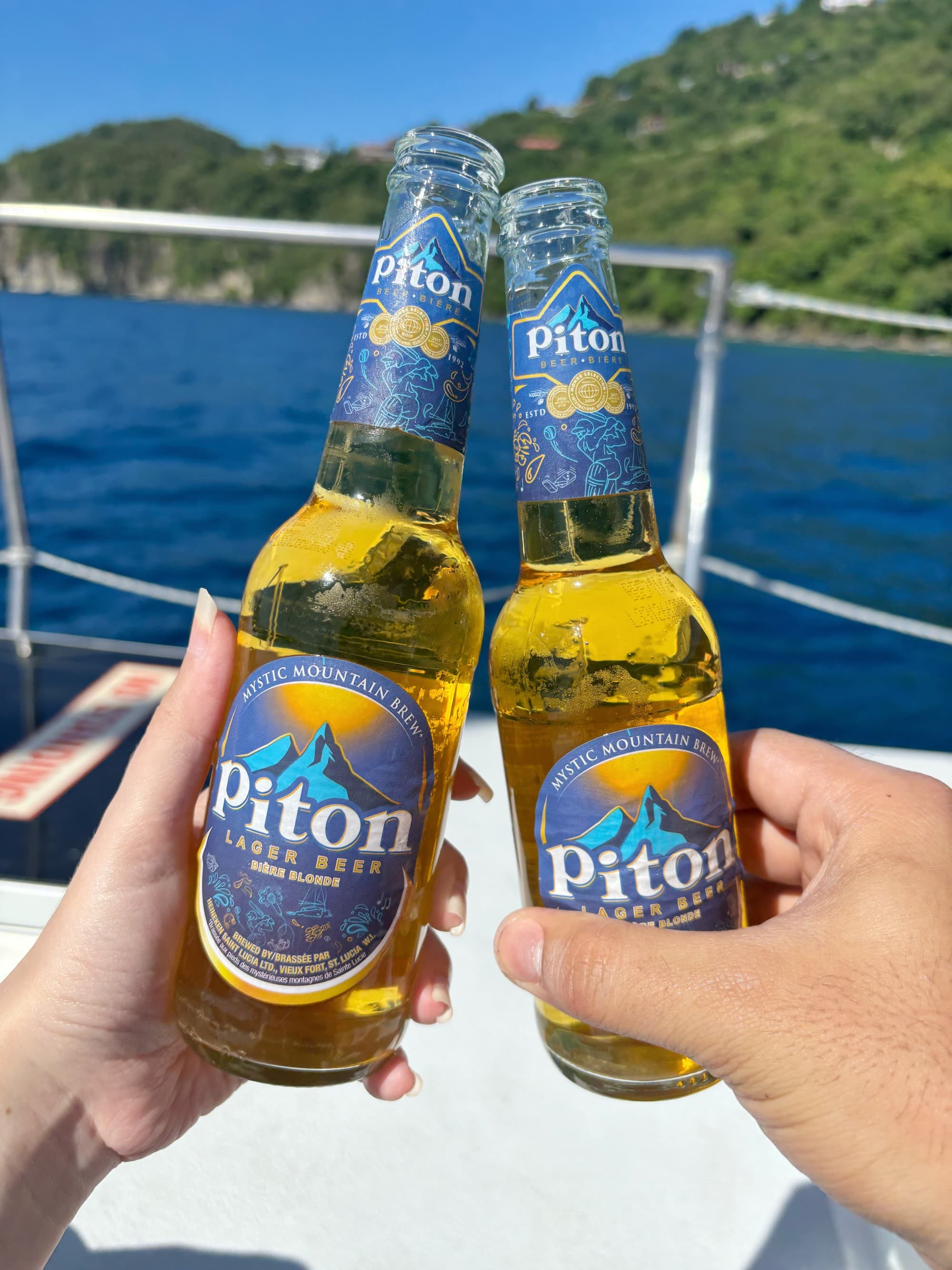 A couple clink beer bottles aboard a boat rounding the shore on a clear day.