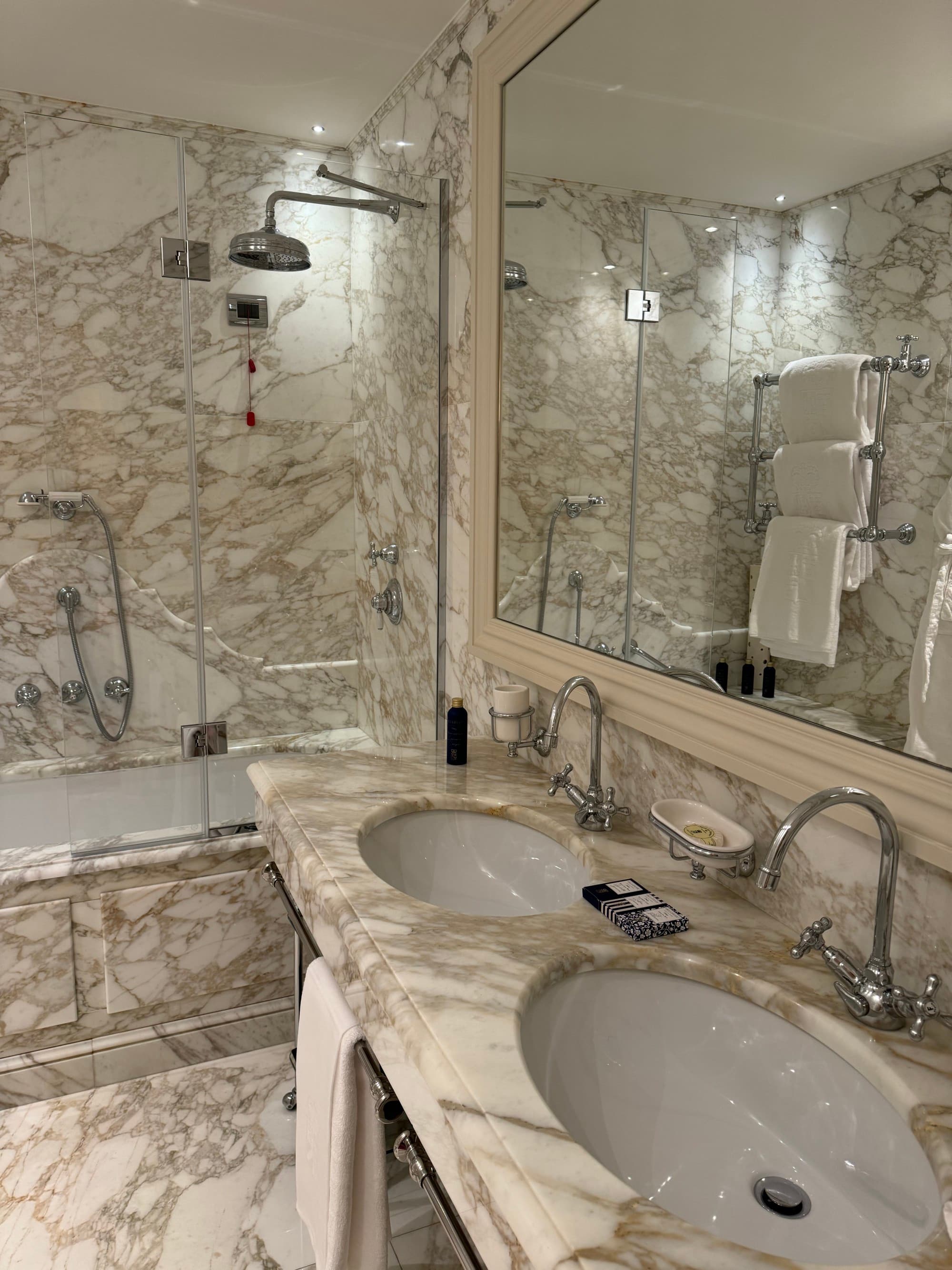 The hotel bathroom is covered in floor to ceiling marble with deep double sinks and a tub.