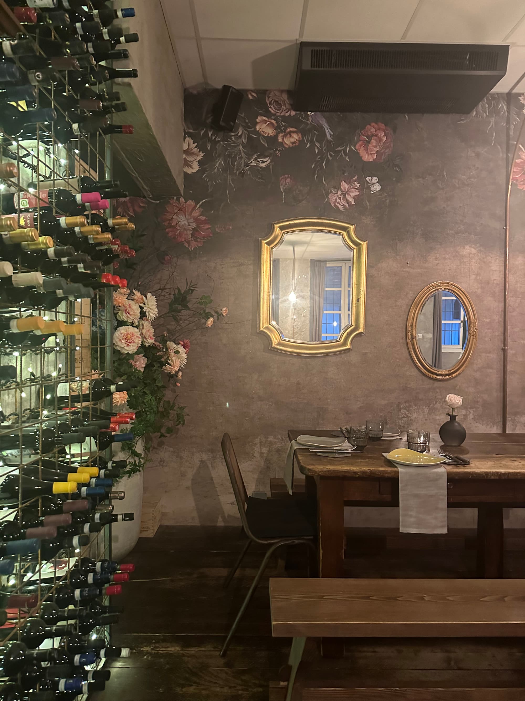 A view of a serene wine bar, shelves stacked with wine bottles, a mirror on the wall and seating.  Osteria interior - Georgia Goddard