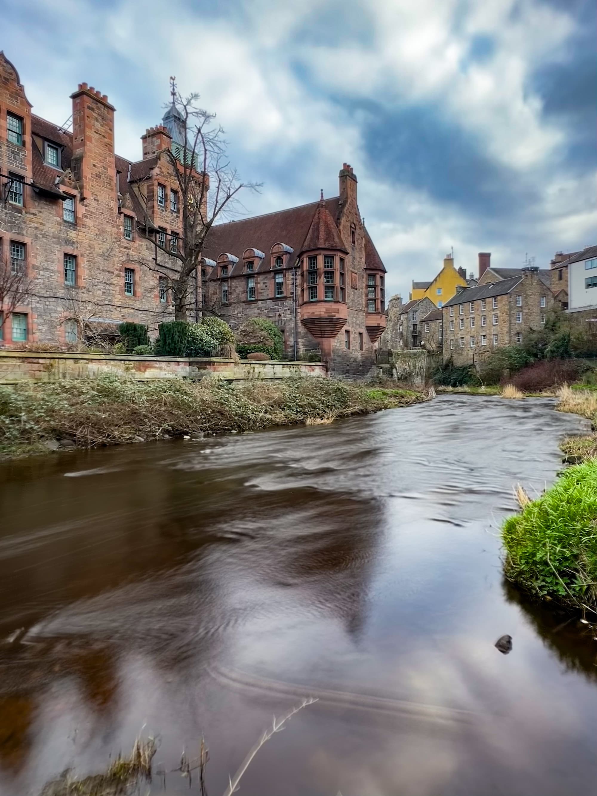 Dean's Village in Edinburgh has a stream running behind brick buildings on a clear day dotted with clouds.