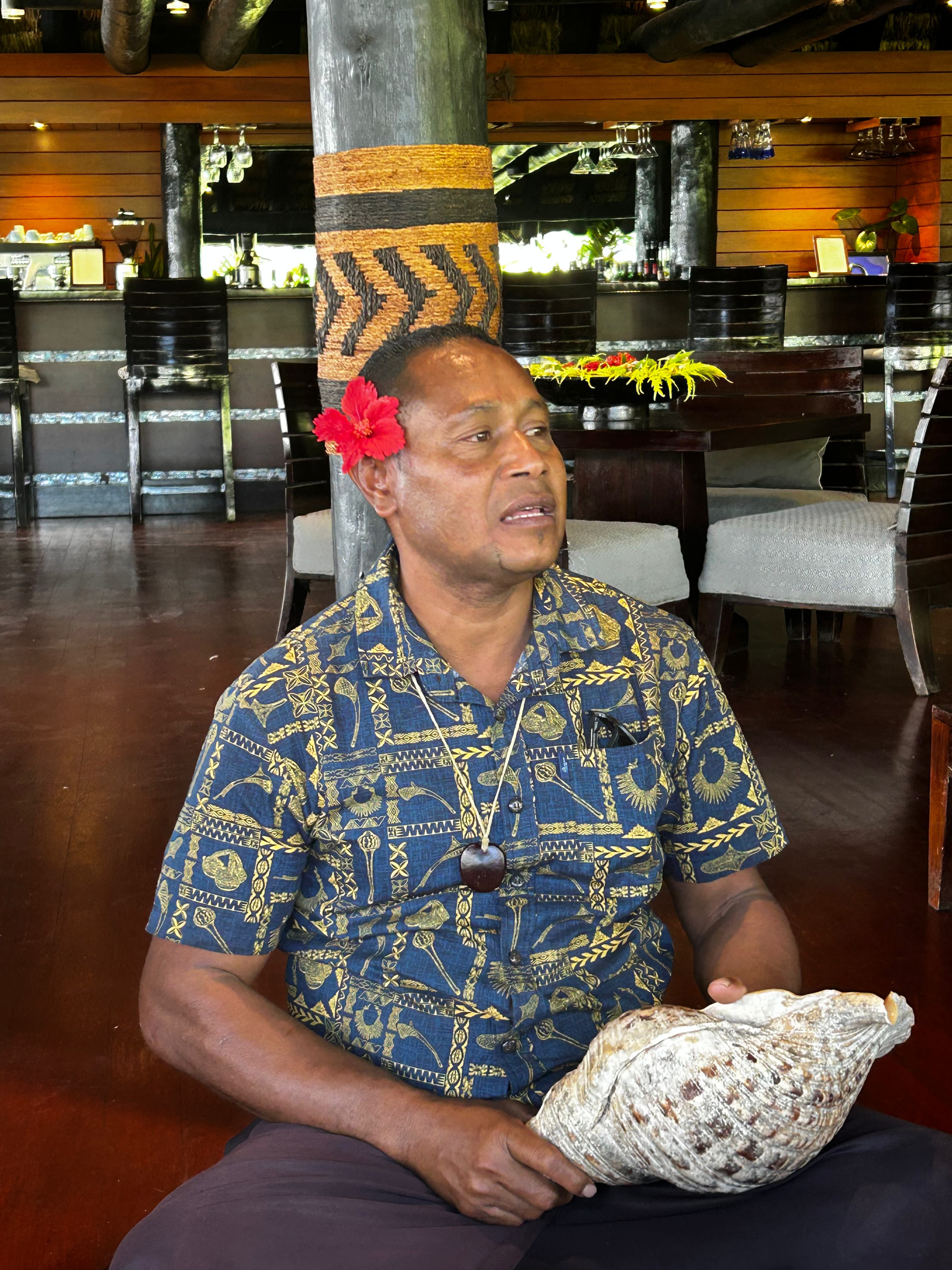 A Fijan man sitting on the floor, holding a large shell with a red flower behind his ear.