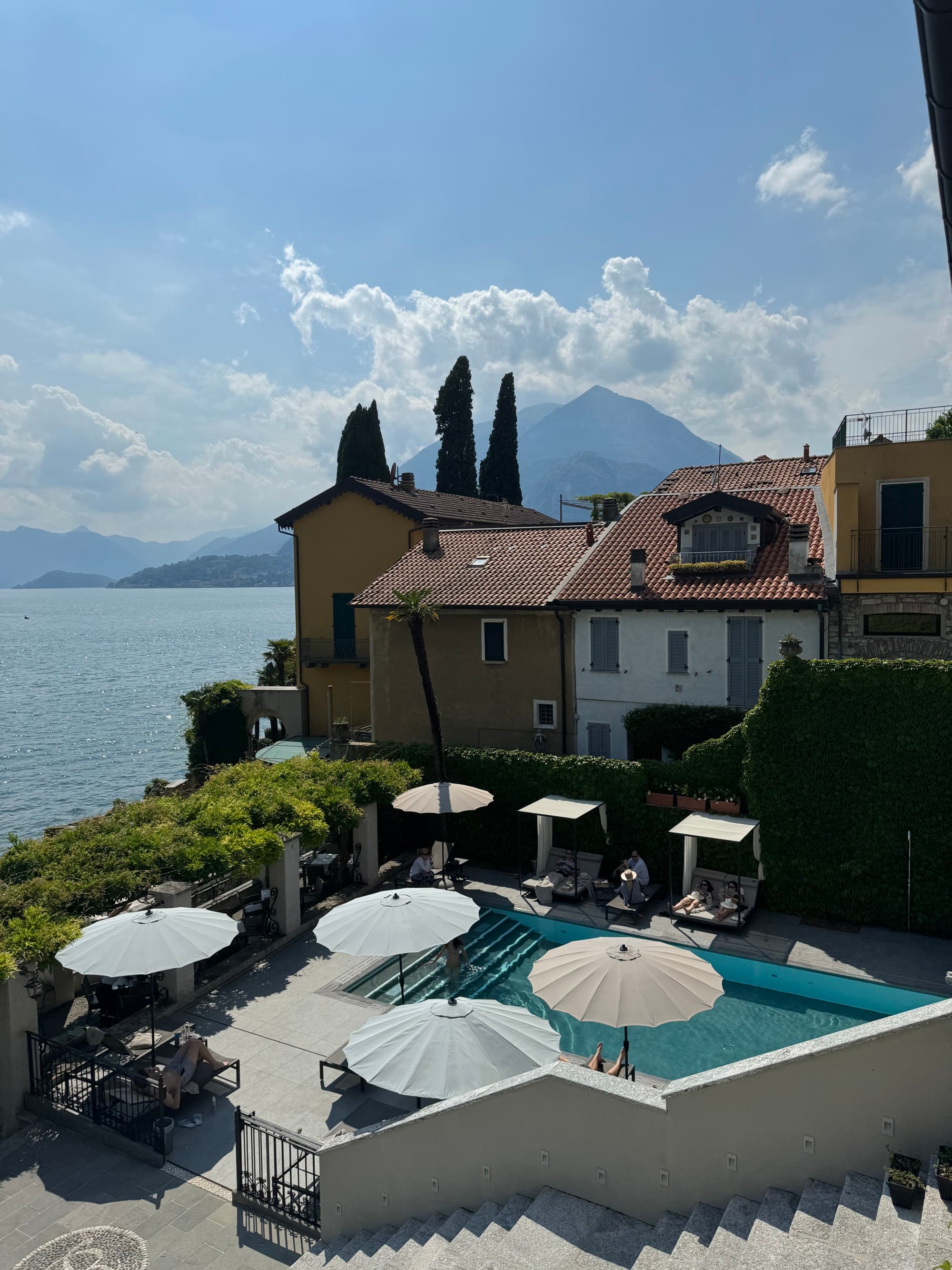 A view of the pool at Hotel Royal Victoria, with white umbrellas, trees and the resort with the lake and mountains in the background.