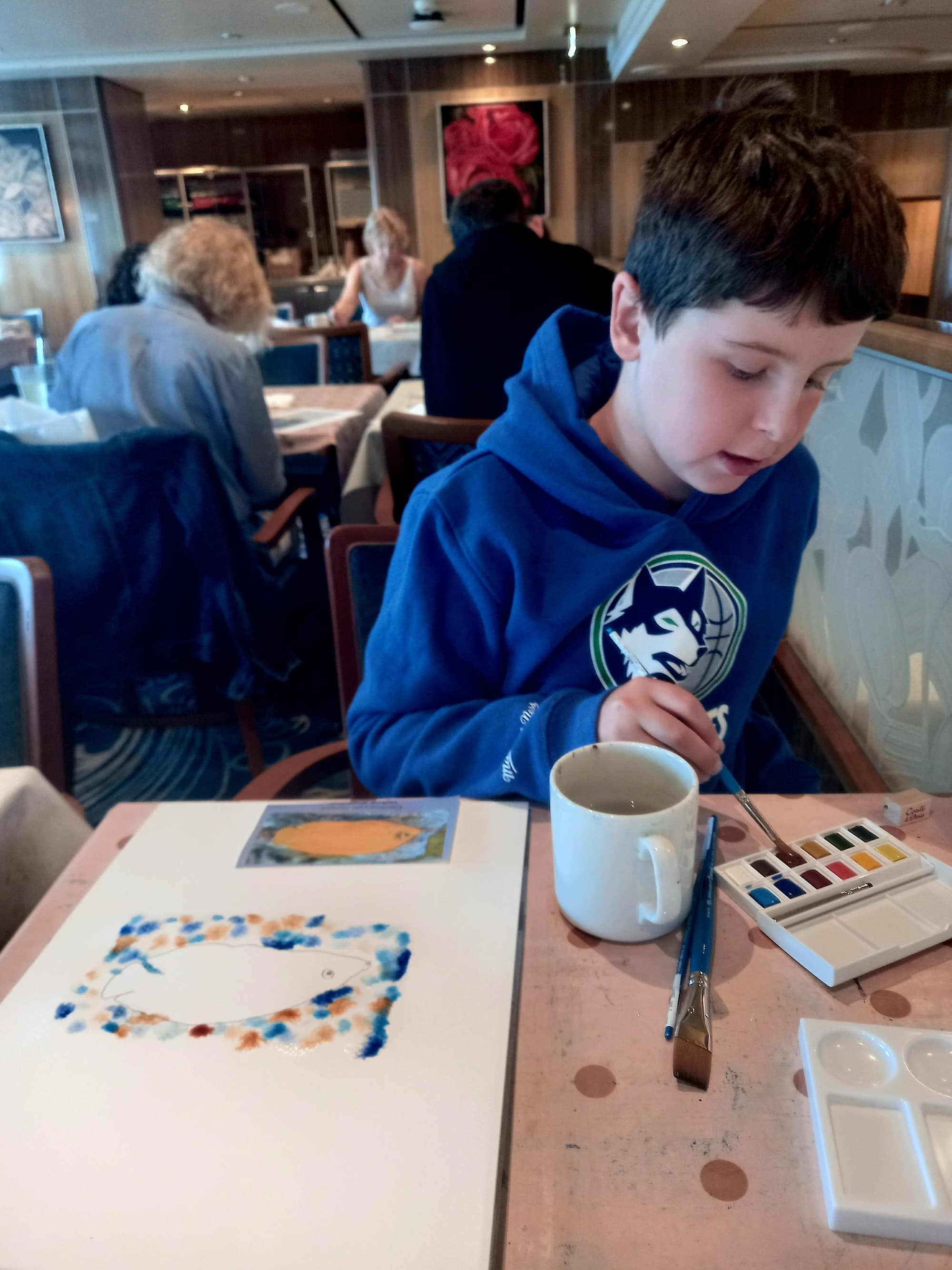 A kid sitting at a table and working on a watercolor painting.