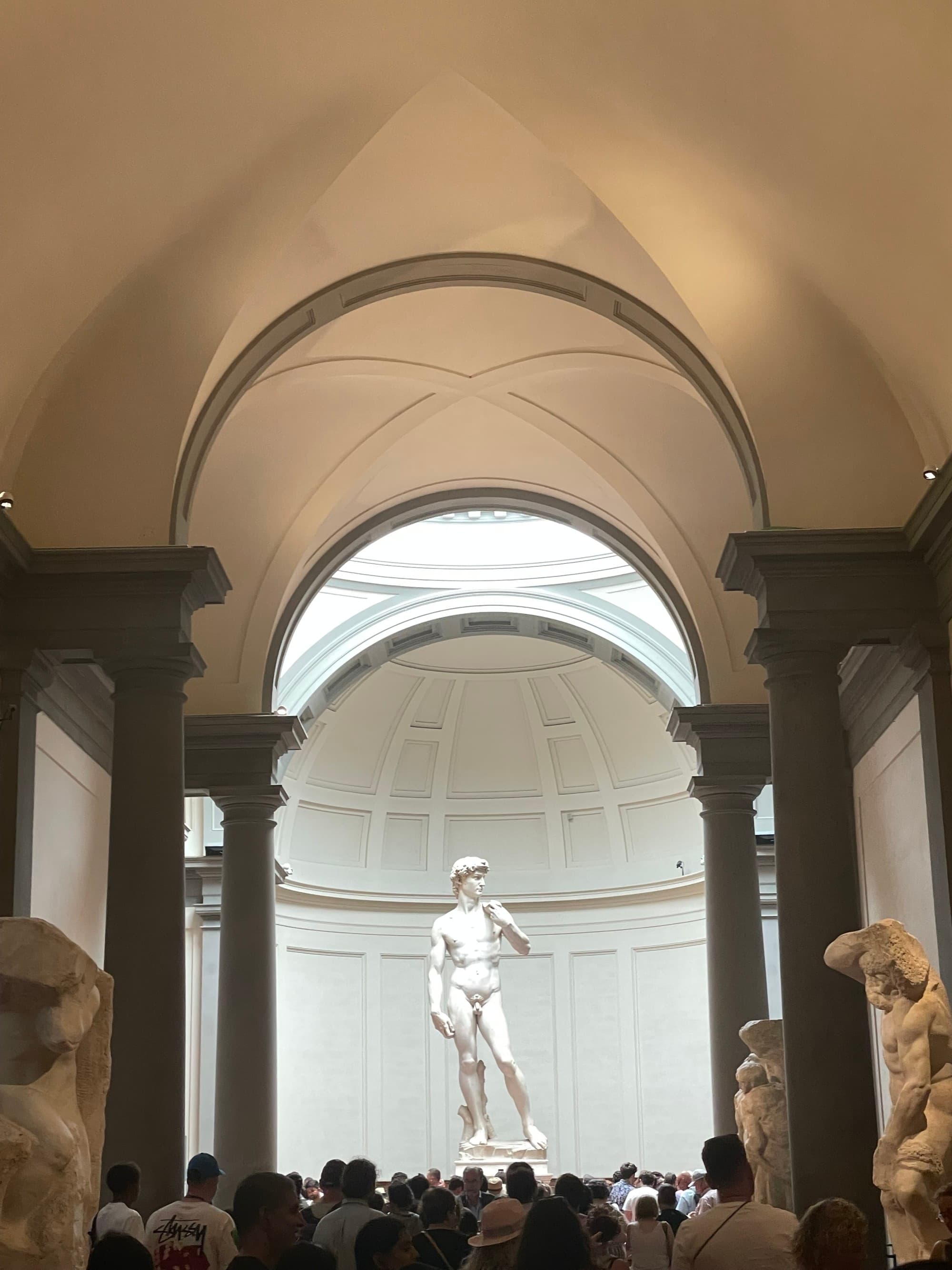 Arched ceilings in a hallway leading to a famous statue of a man in a museum with a large crowd of people around it