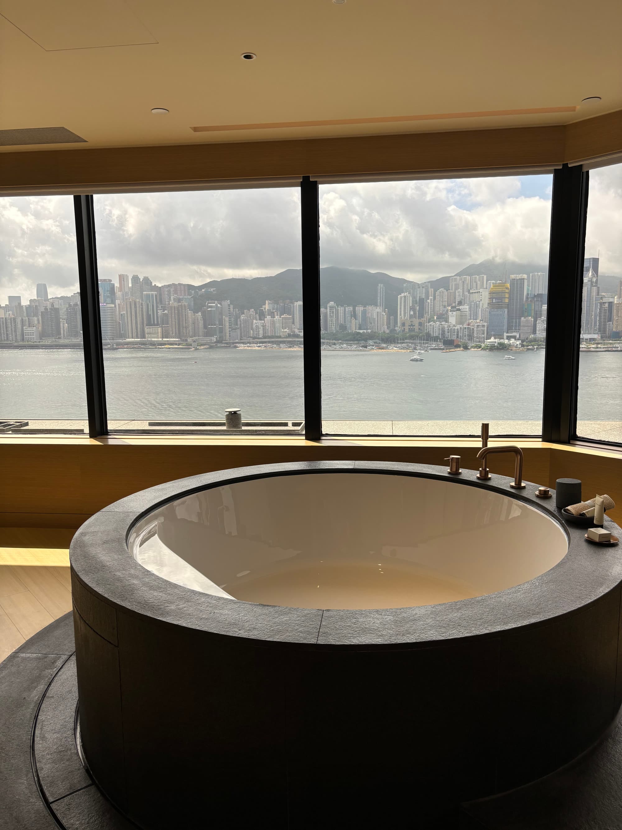 A large, round bath tub in front of large windows offering a view of a body of water with city buildings in the distance