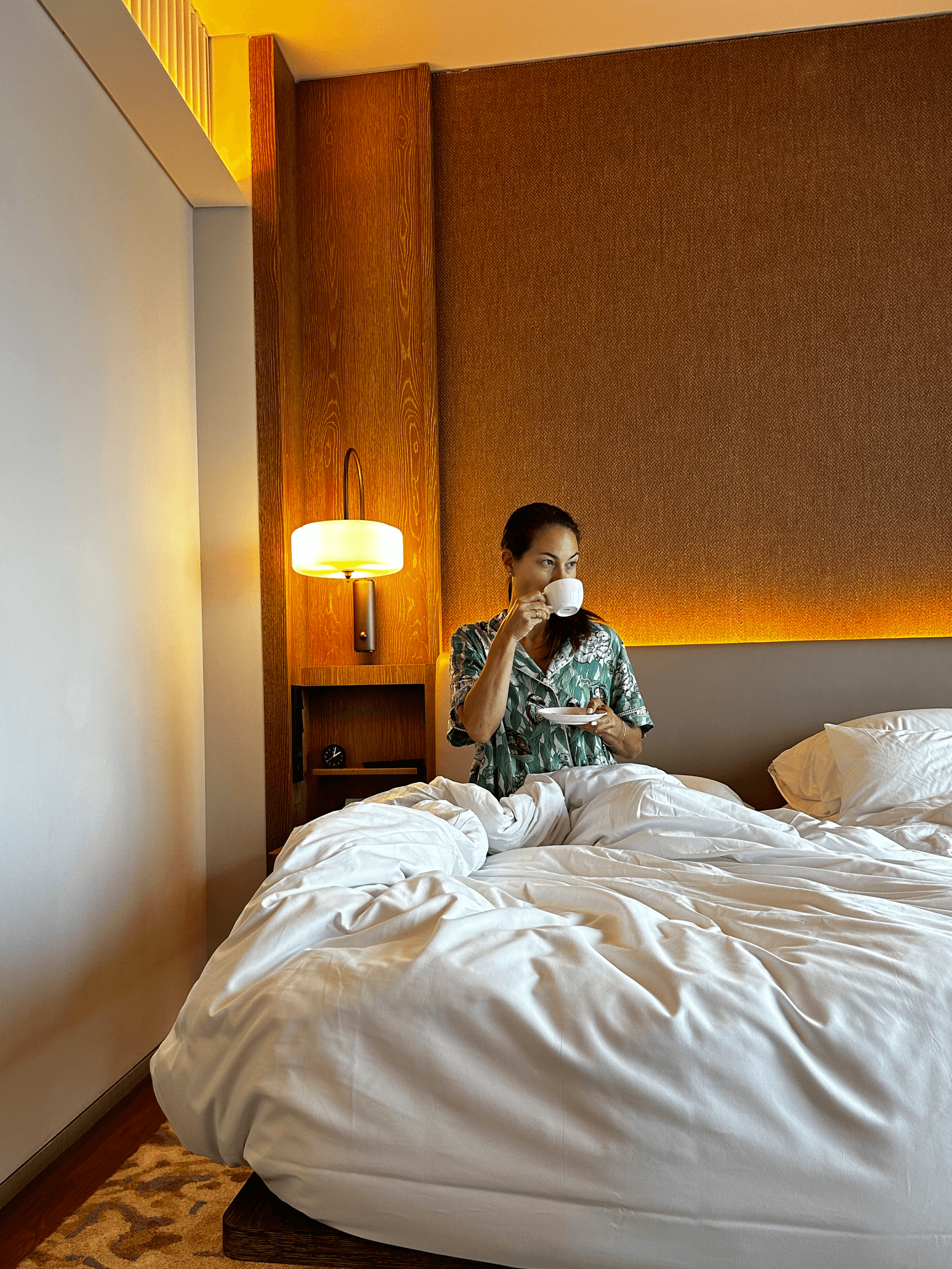 A person in a hotel bed sipping a drink from a white mug