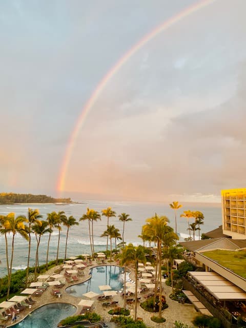 A picture of a beautiful rainbow shining over a bay with a hotel patio, pool, lounge chairs and palm trees in the forefront of the view.