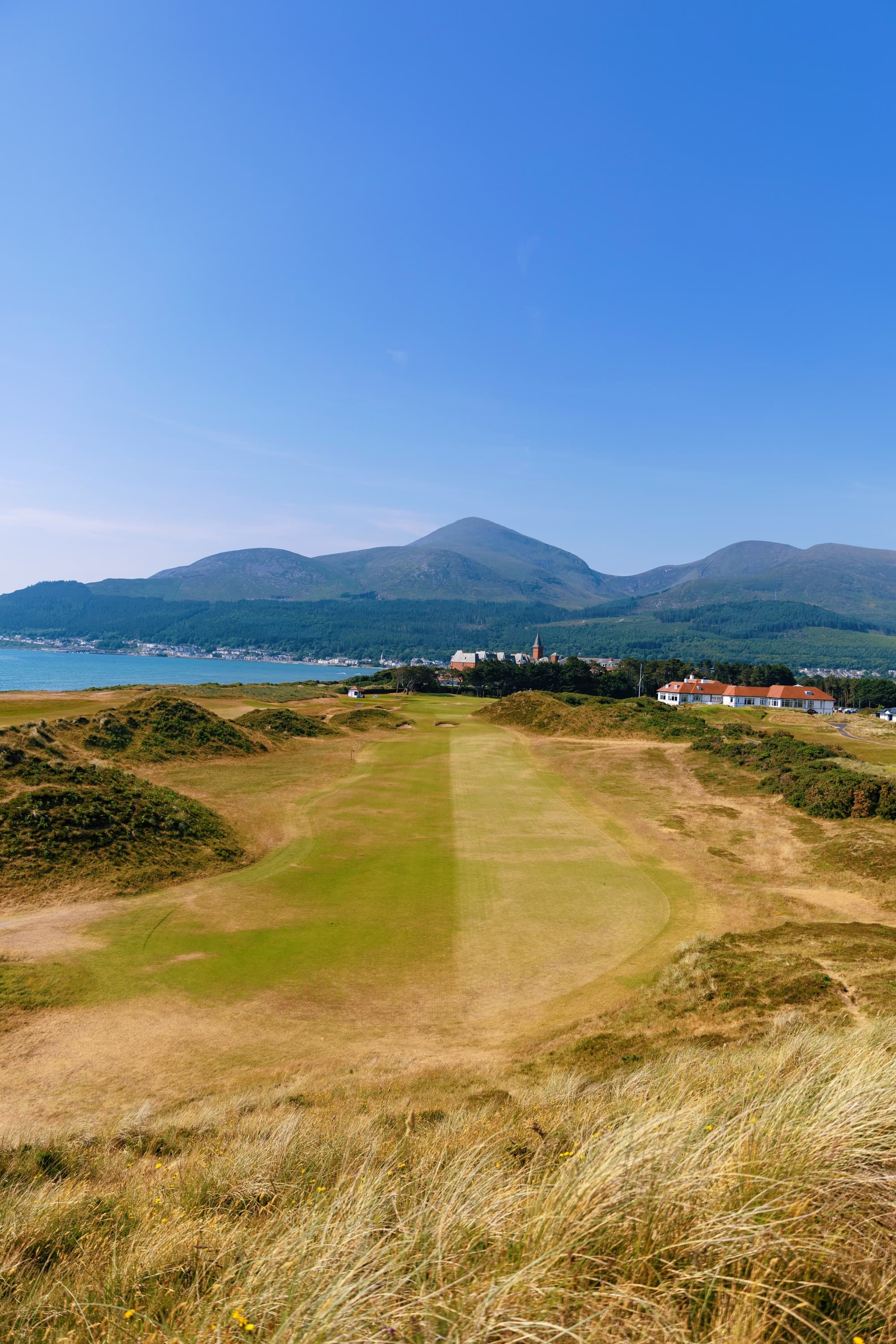 A scenic golf course with undulating terrain, leading towards a mountain range under a clear blue sky.