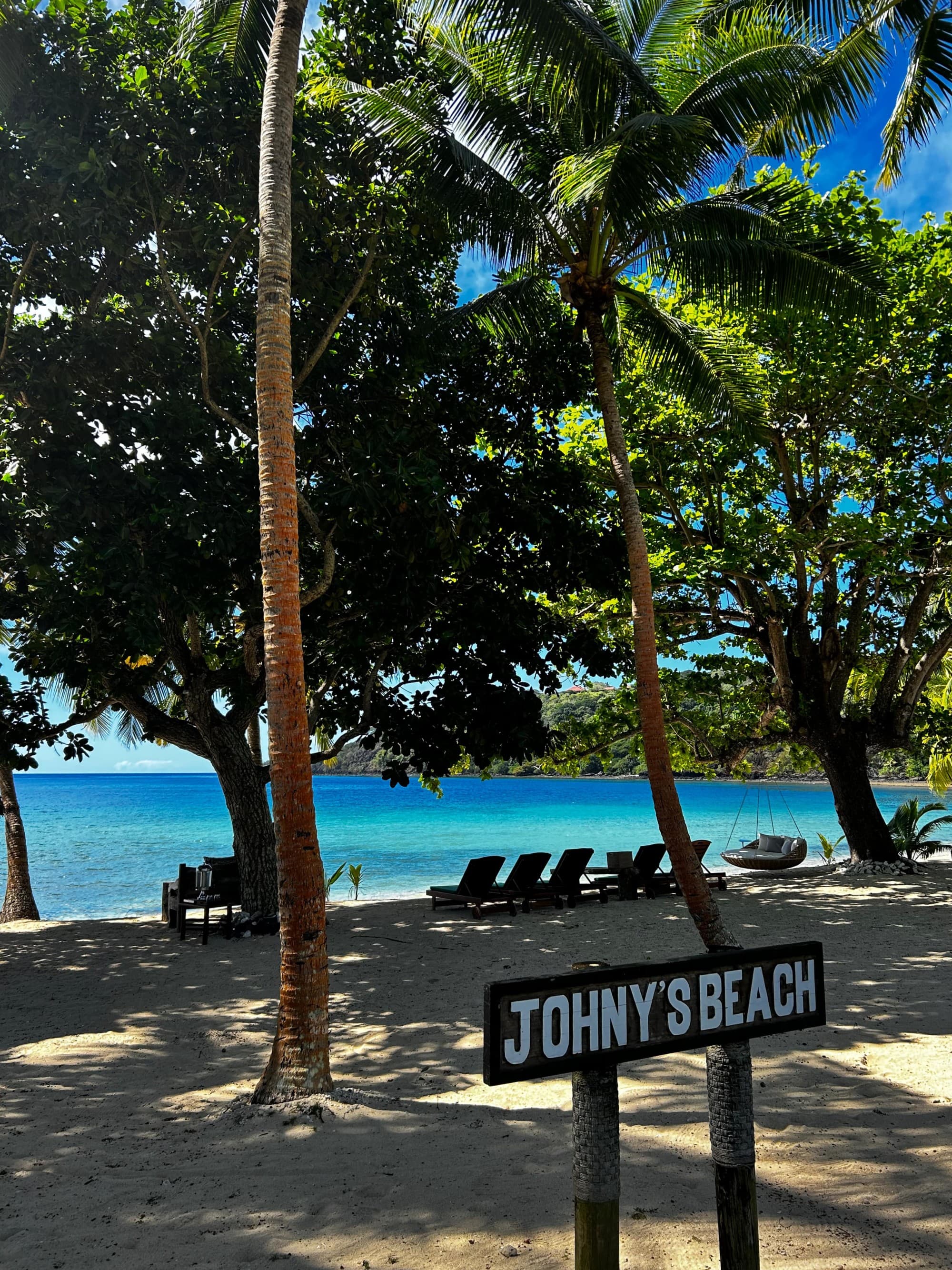 A beach during the daytime with a wooden sign reading "Johny's Beach" in front of a group of trees