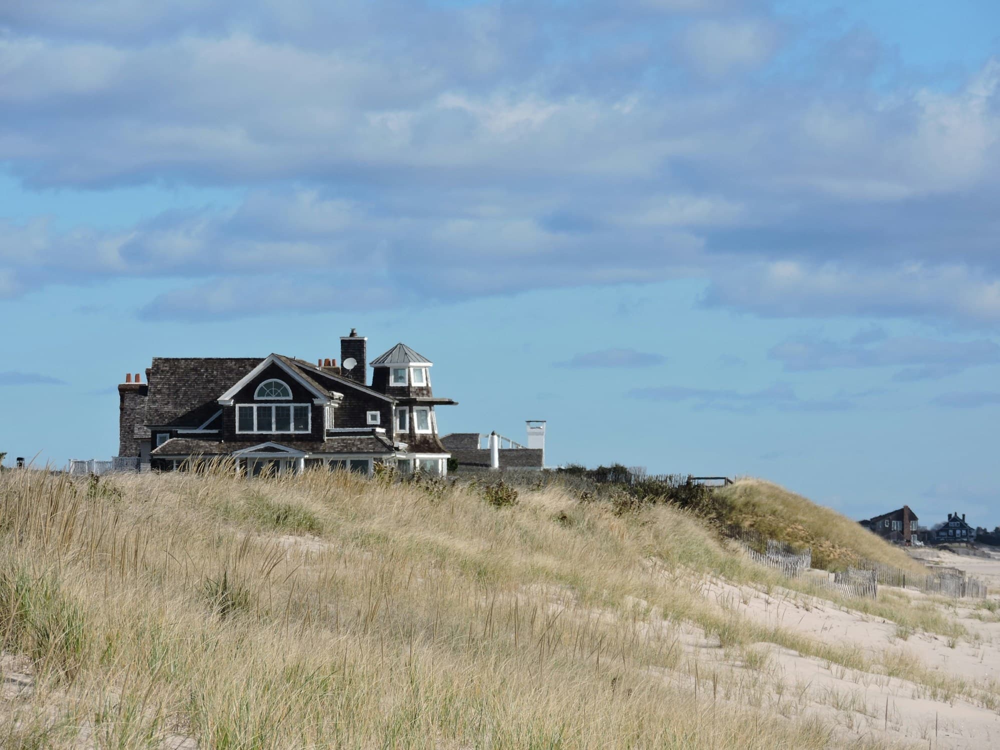 Hampton oceanfront house on a grassy hill as sand and ocean waves swirl below on a cloudy day.