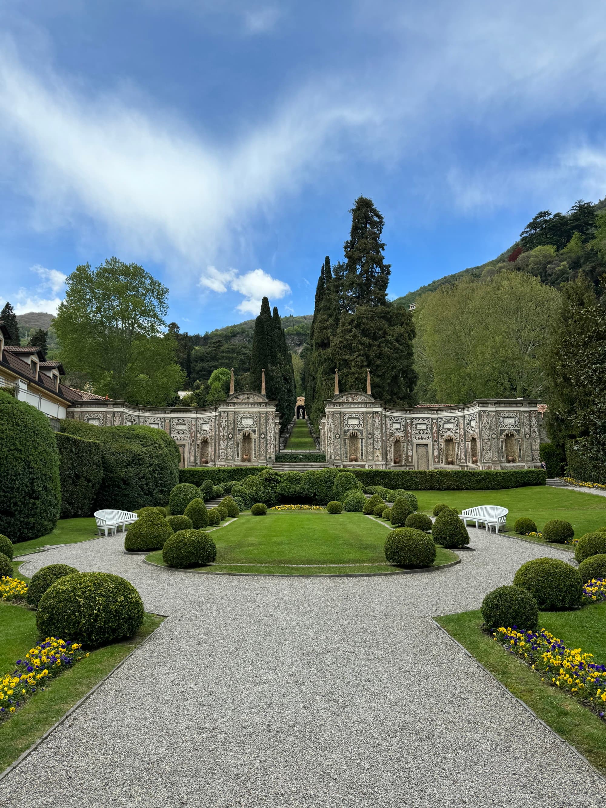 Manicured gardens surround a cobbled path lined with evergreen trees on a clear day dotted with clouds.