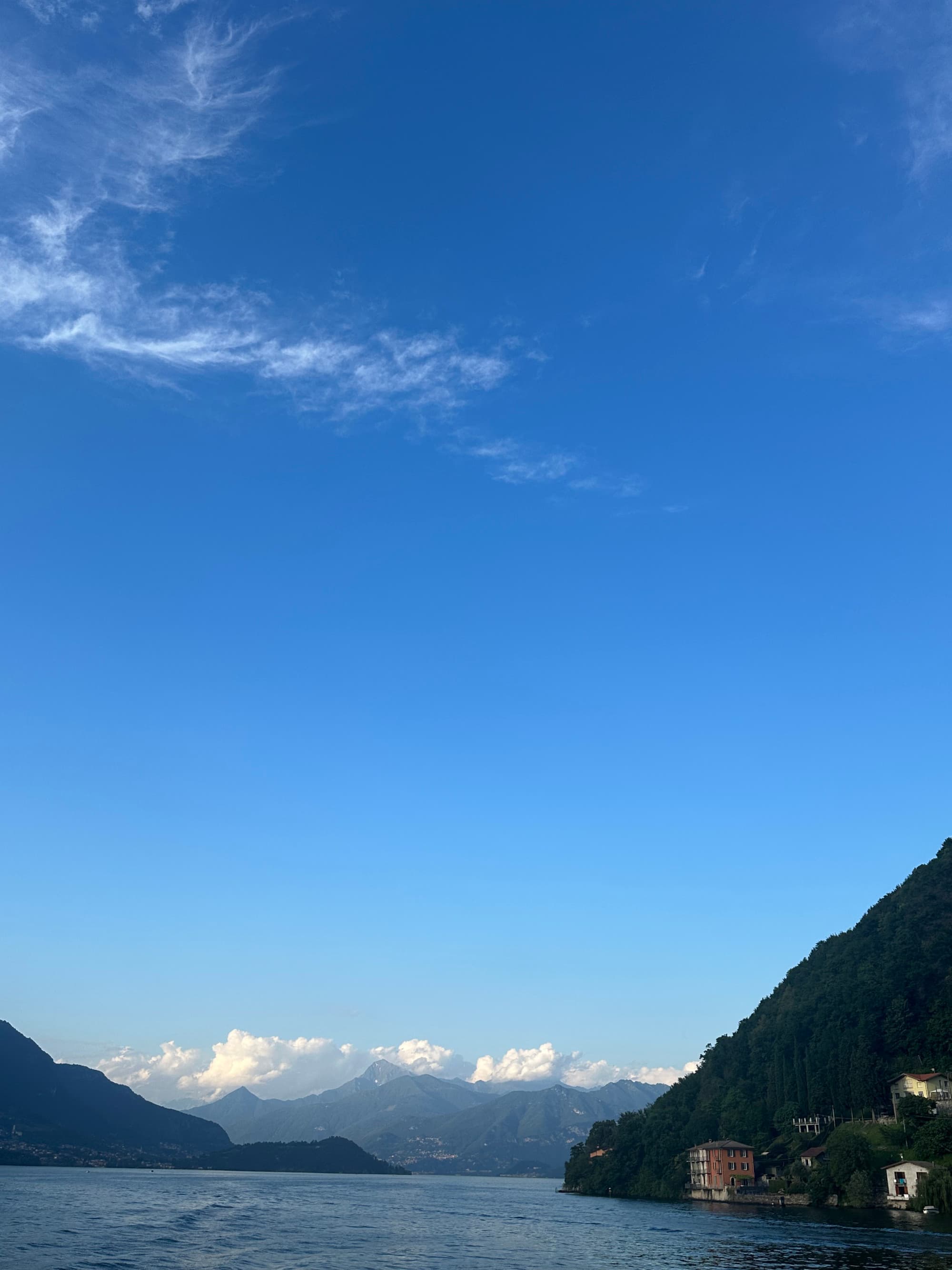 A view from the ferry on the lale with mountains, low puffy white clouds and a big blue sky during the day.