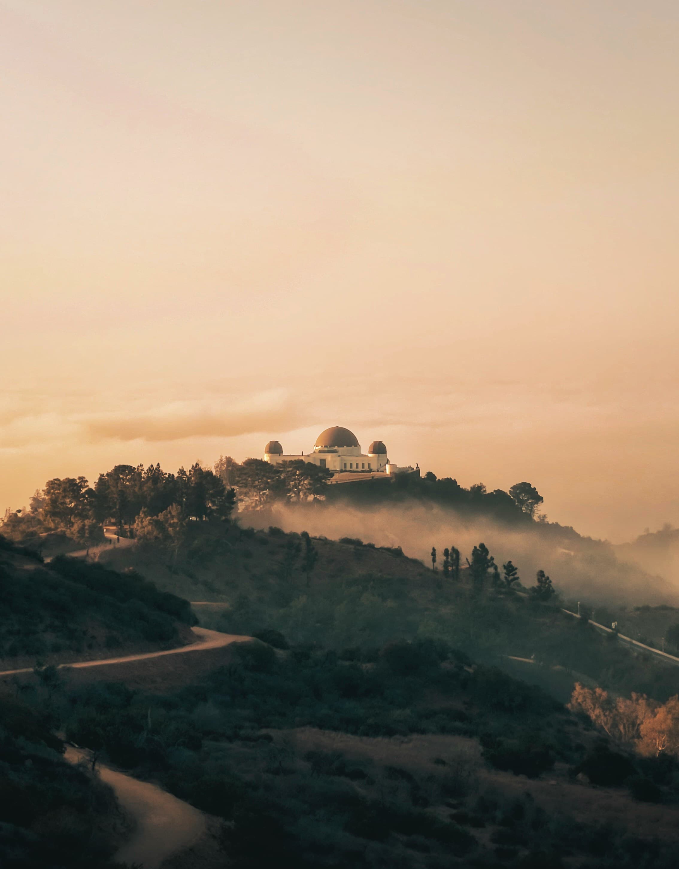 A view from one of the hotels near Griffith Observatory, featuring a large white building with a domed roof, on top of a hill covered in trees at sunset.