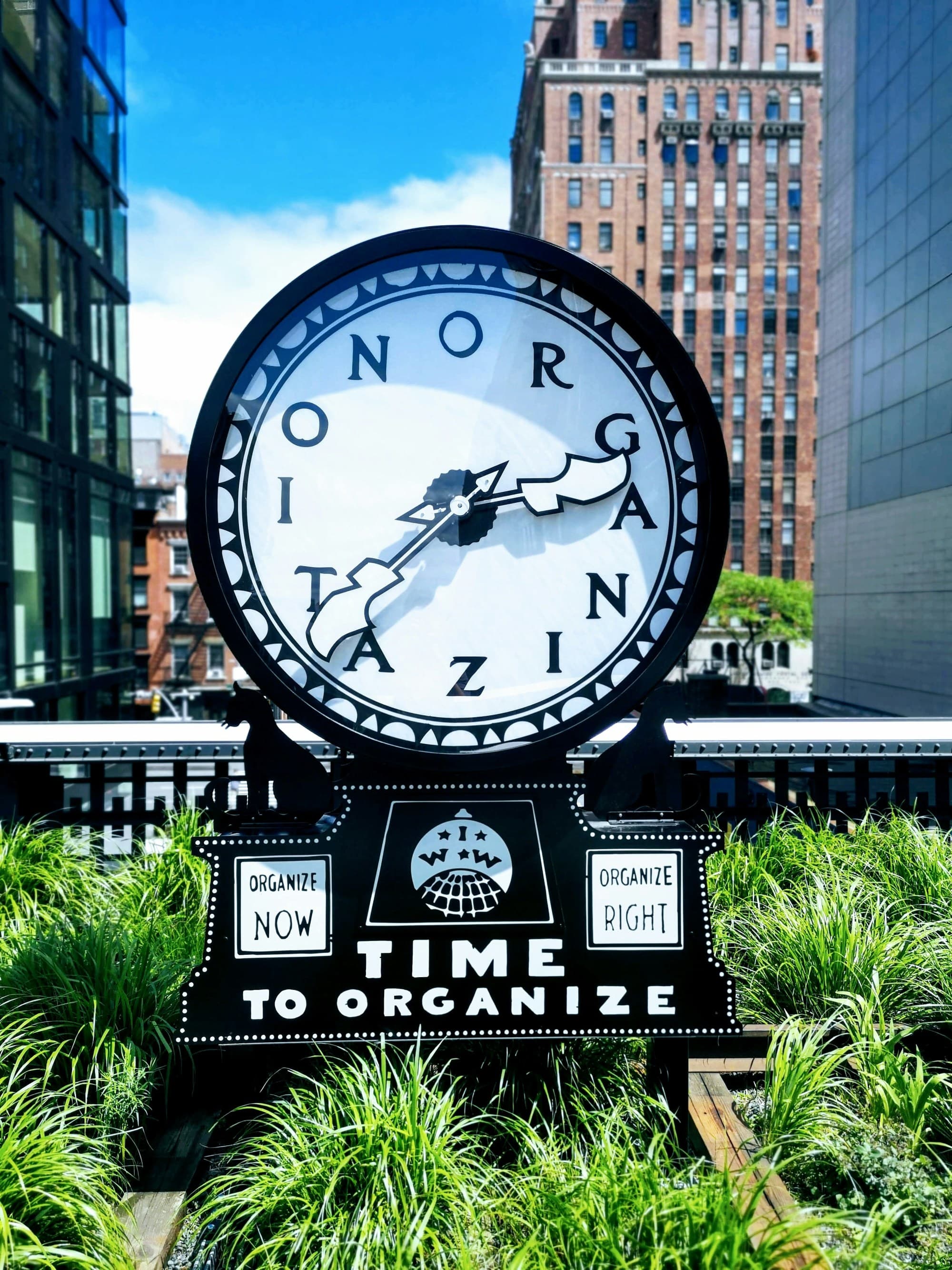 Clock sculpture that says "time to organize" amidst city buildings and greenery