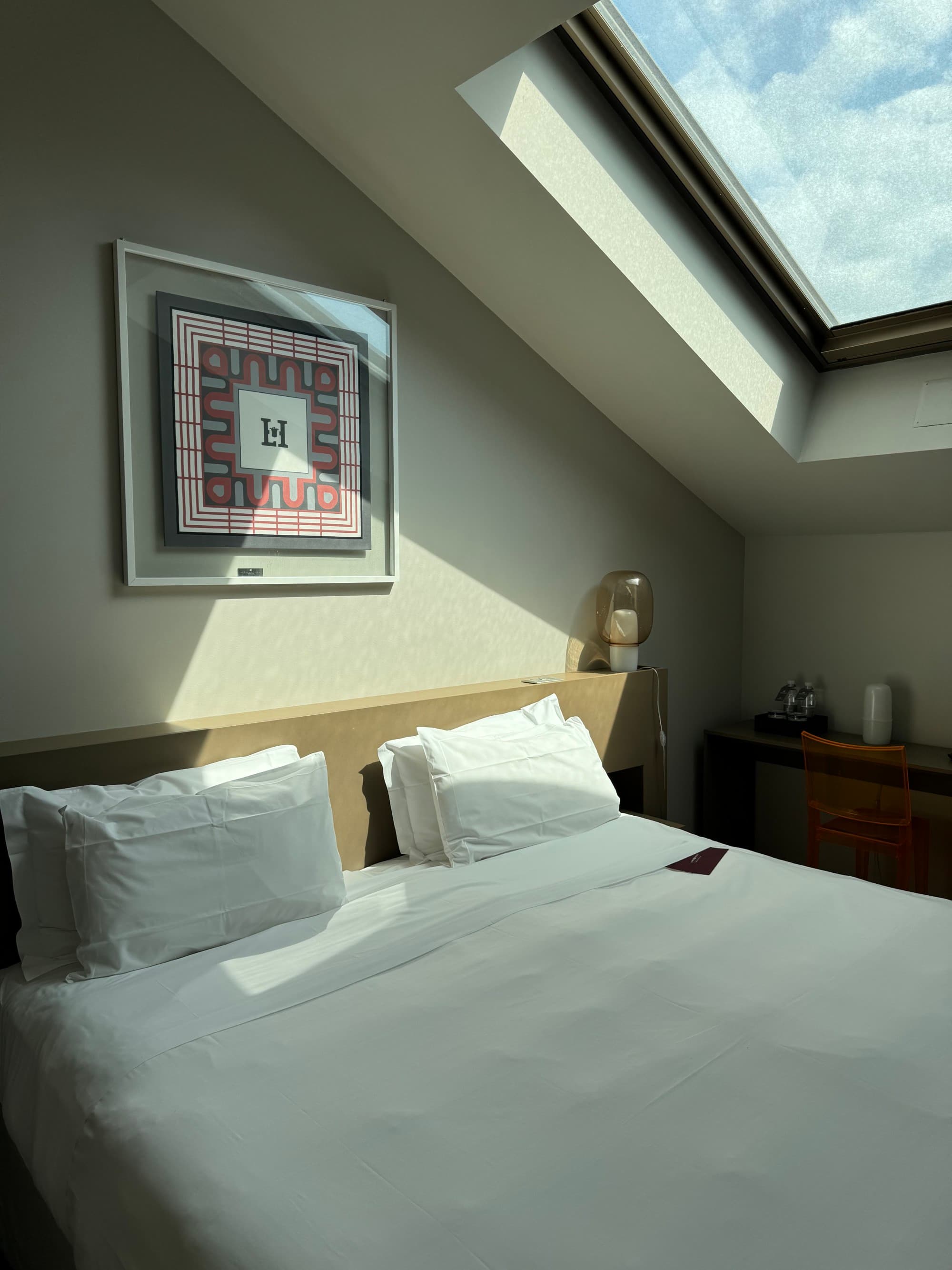 A photo of a white linen bed underneath an A-frame roof with a window letting light in from it onto the bed.