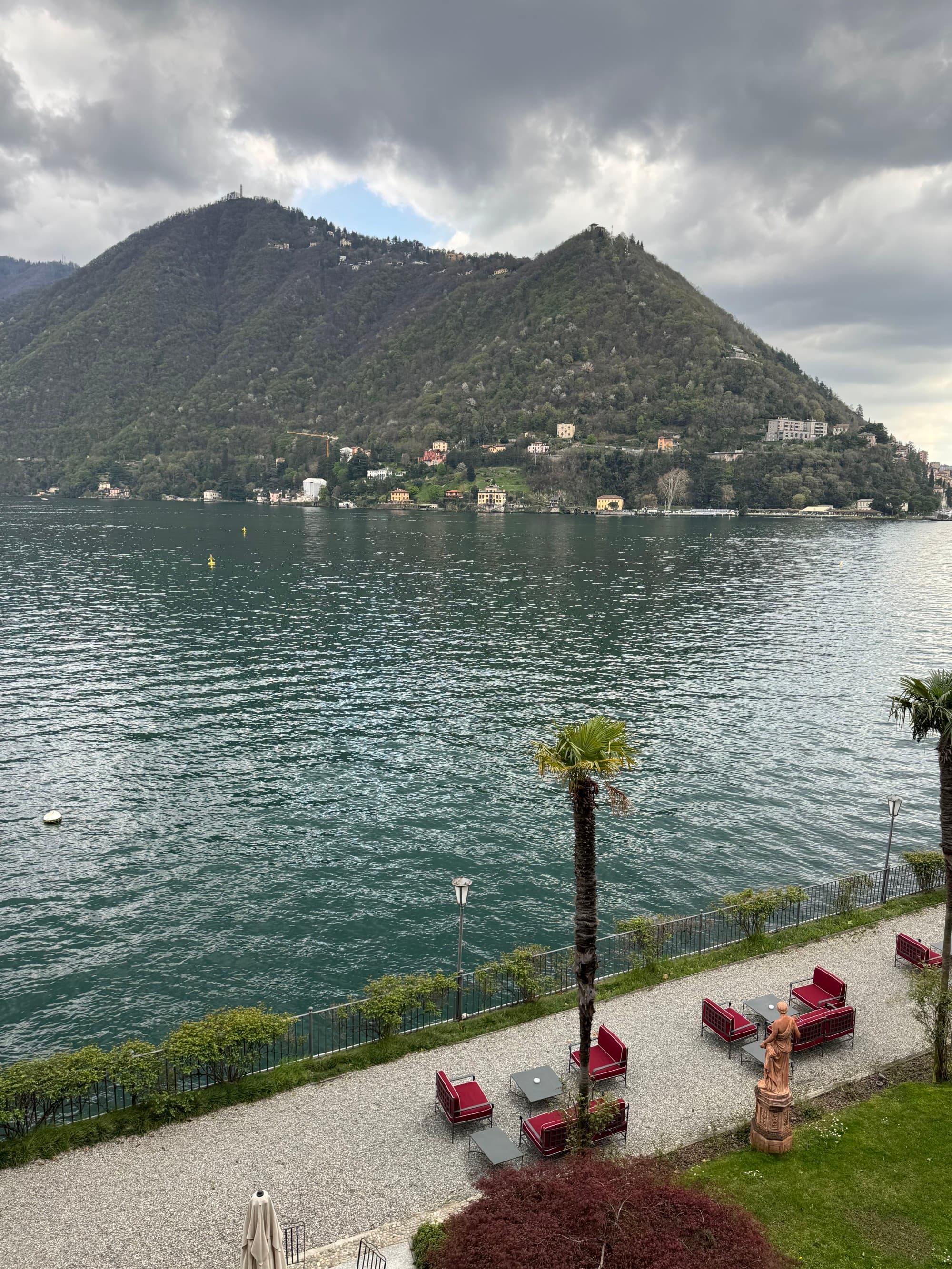 A view of Lake Como and its walkways with lounge chairs below. A large mountain stands on the opposite side of the lake.