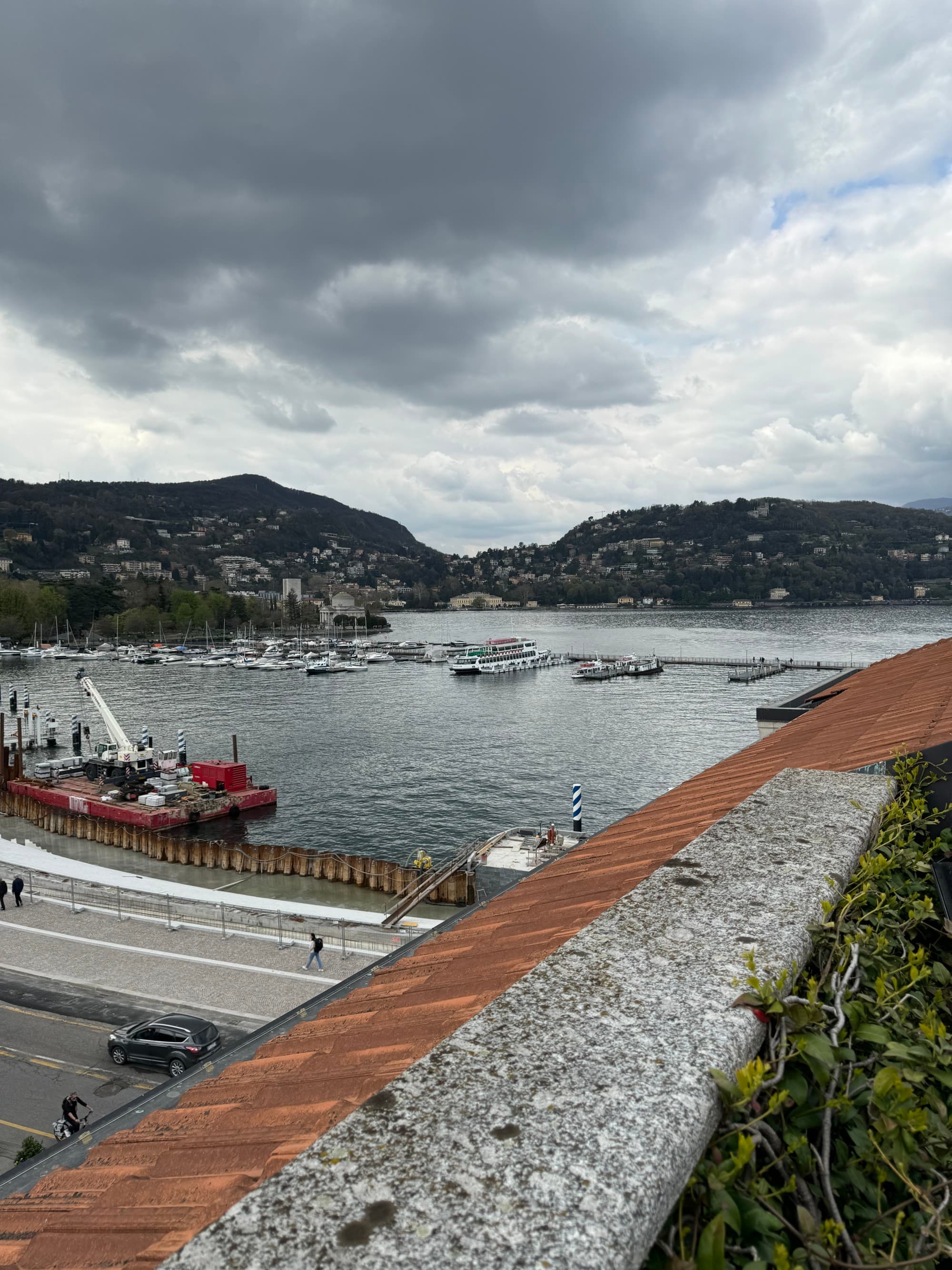 A view of a small harbor in Lake Como from an overlook point. There are green mountains in the distance and the sky is cloudy and gloomy.