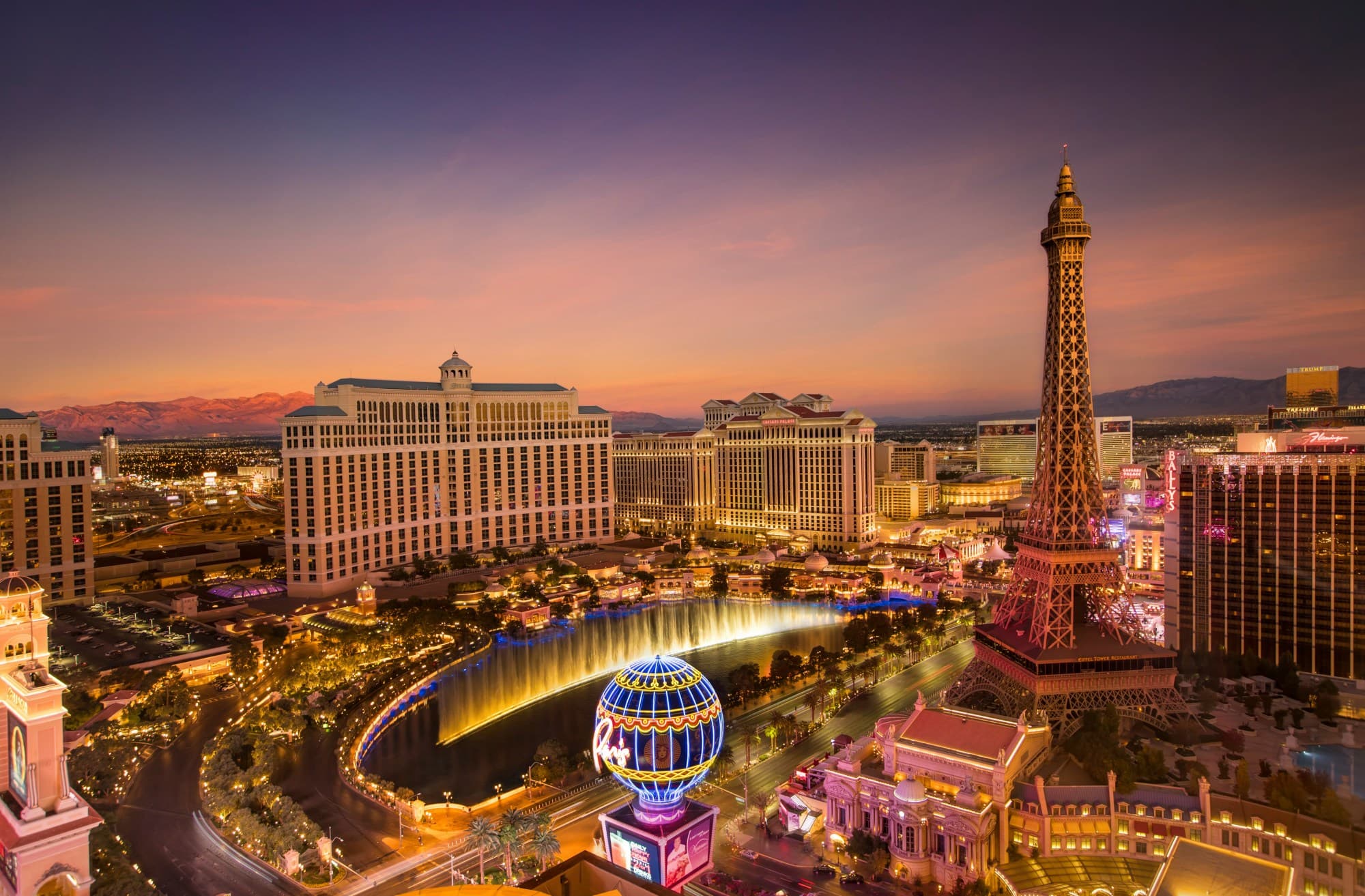 An aerial view from the Encore Tower Suites, highlighting the Bellagio Hotel and fountain, mock Eiffel Tower and other Las Vegas buildings, some lit up before a dusk sky.