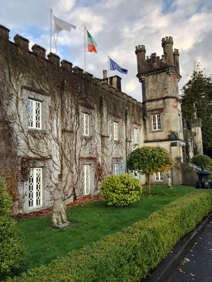 A view of a stone castle's exterior with three flags perched on top, with a green lawn and trees surrounding it on a sunny day.