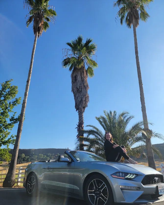 Picture of Ashlyn sitting on the hood of a 2020 Ford Mustang car on a sunny day