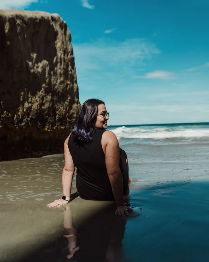 Rear view of women sitting on the sand on a beach in a black top with a large rock formation in view