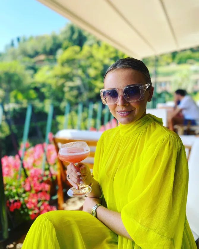 Travel advisor Edwina in a yellow dress and sunglasses holding a cocktail with a beautiful garden view behind