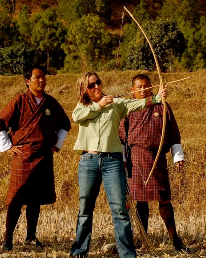 Travel advisor Allison shooting a bow and arrow surrounded by greenery with two men in brown robes watching on