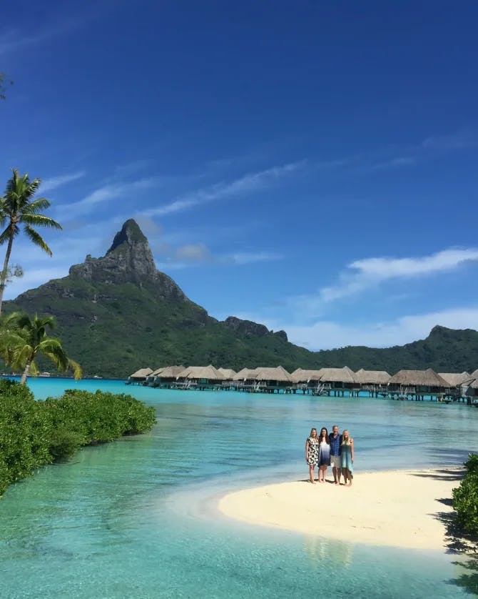 A picture of four people standing on a small sandy inlet surrounded by turquoise water with overwater bungalows in the background