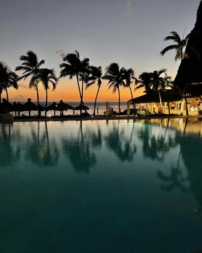 Beautiful view of a pool surrounded by palm trees and cabanas on the beach at sunset